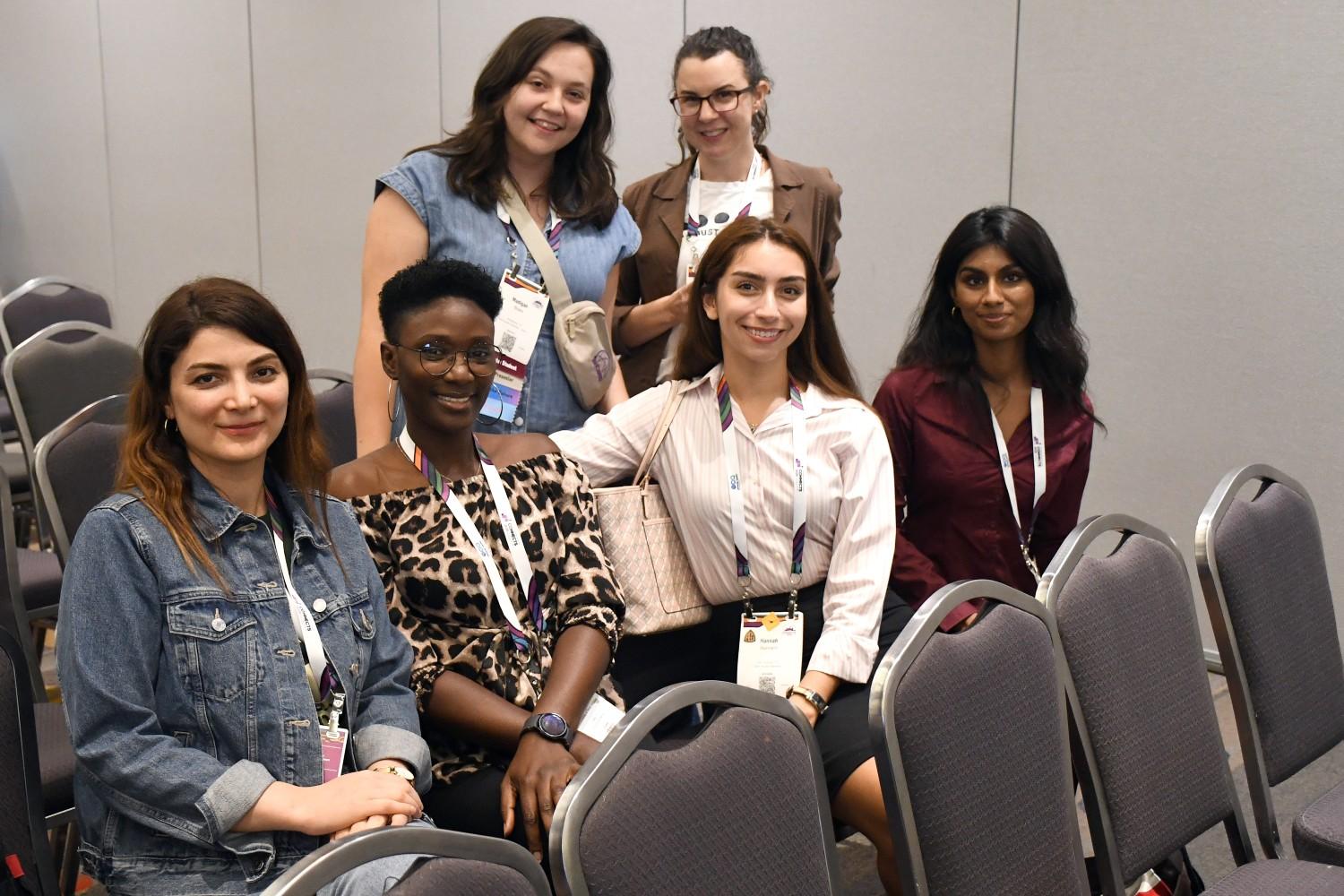Six women smiling at a conference, sitting and standing among gray chairs. They wear lanyards and casual business attire, conveying a friendly atmosphere.