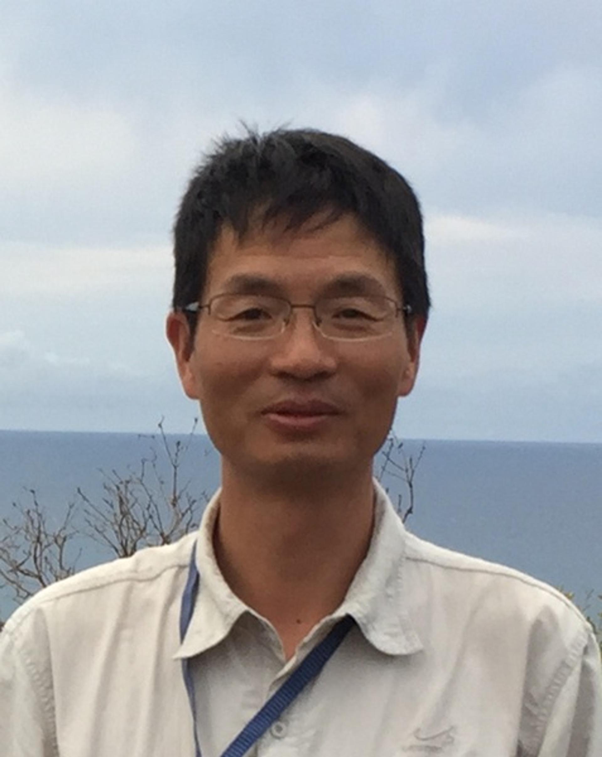 Zhen Yan in a light colored shirt with water, sky, and plants in the background.