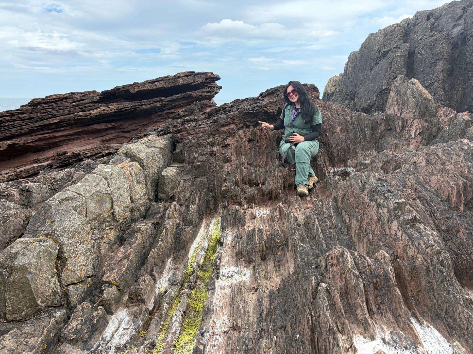 A woman with long brown hair and sunglasses sits on craggy rocks and points and smiles.