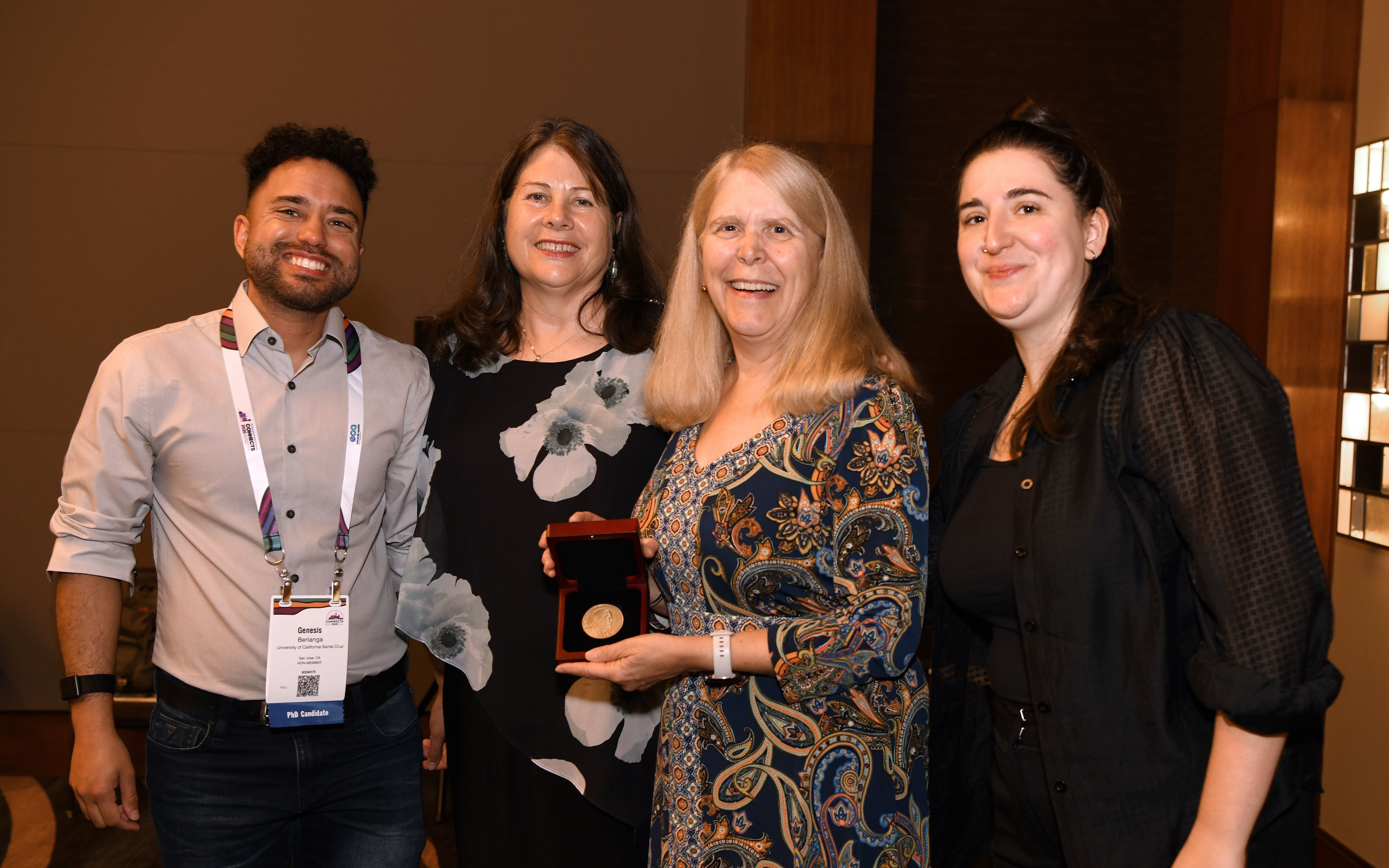 Four Connects 2025 attendees pose together at a conference event, with one woman holding an award medal in an open presentation case while the group smiles for the photo in a warmly lit venue hallway.