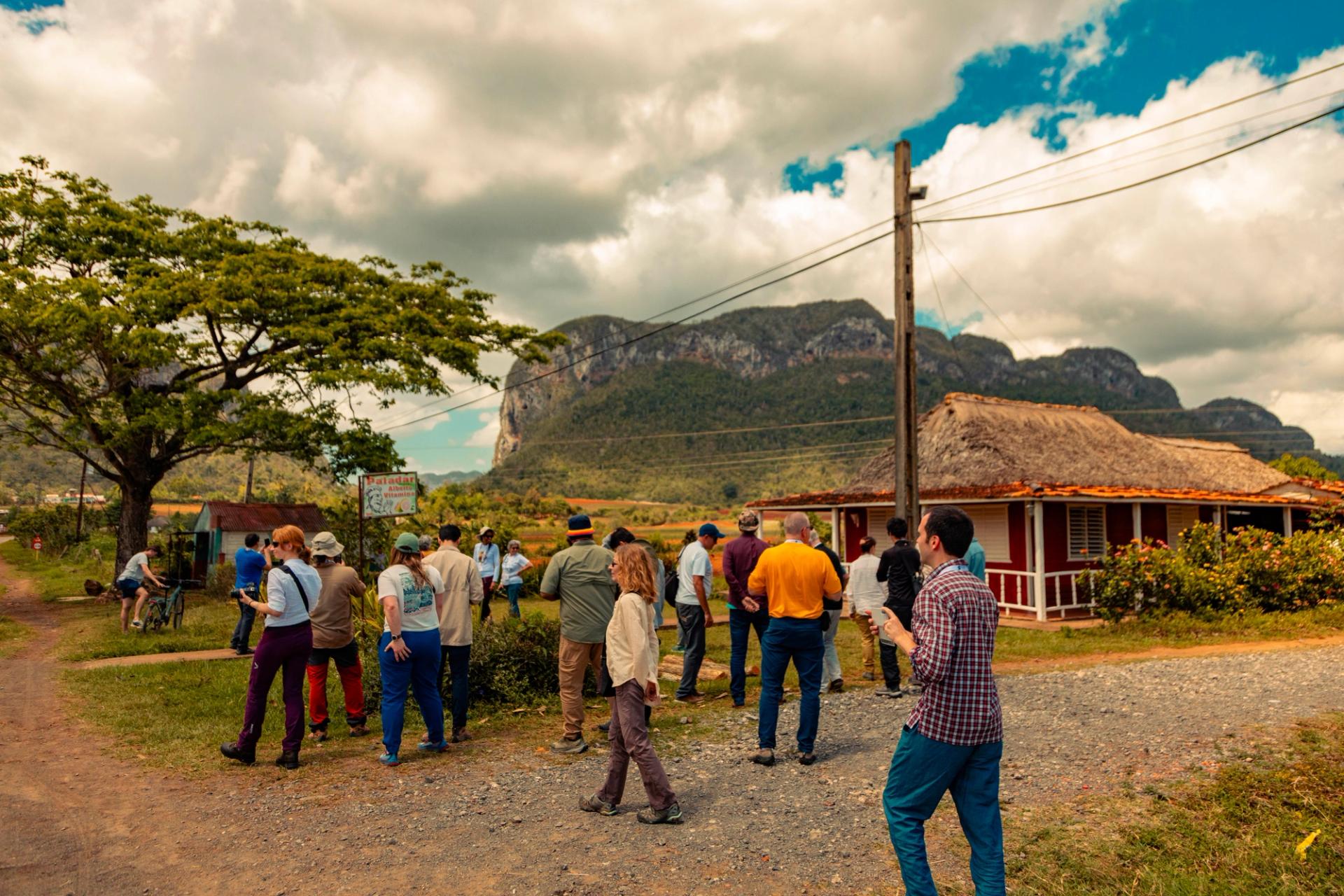 A group of GSA Thompson Field Forum 2025 participants gather on a gravel road in the rural village of Viñales, Cuba, with a traditional thatched-roof building, lush vegetation, and dramatic limestone mogote formations rising under a dramatic cloudy sky in the background.