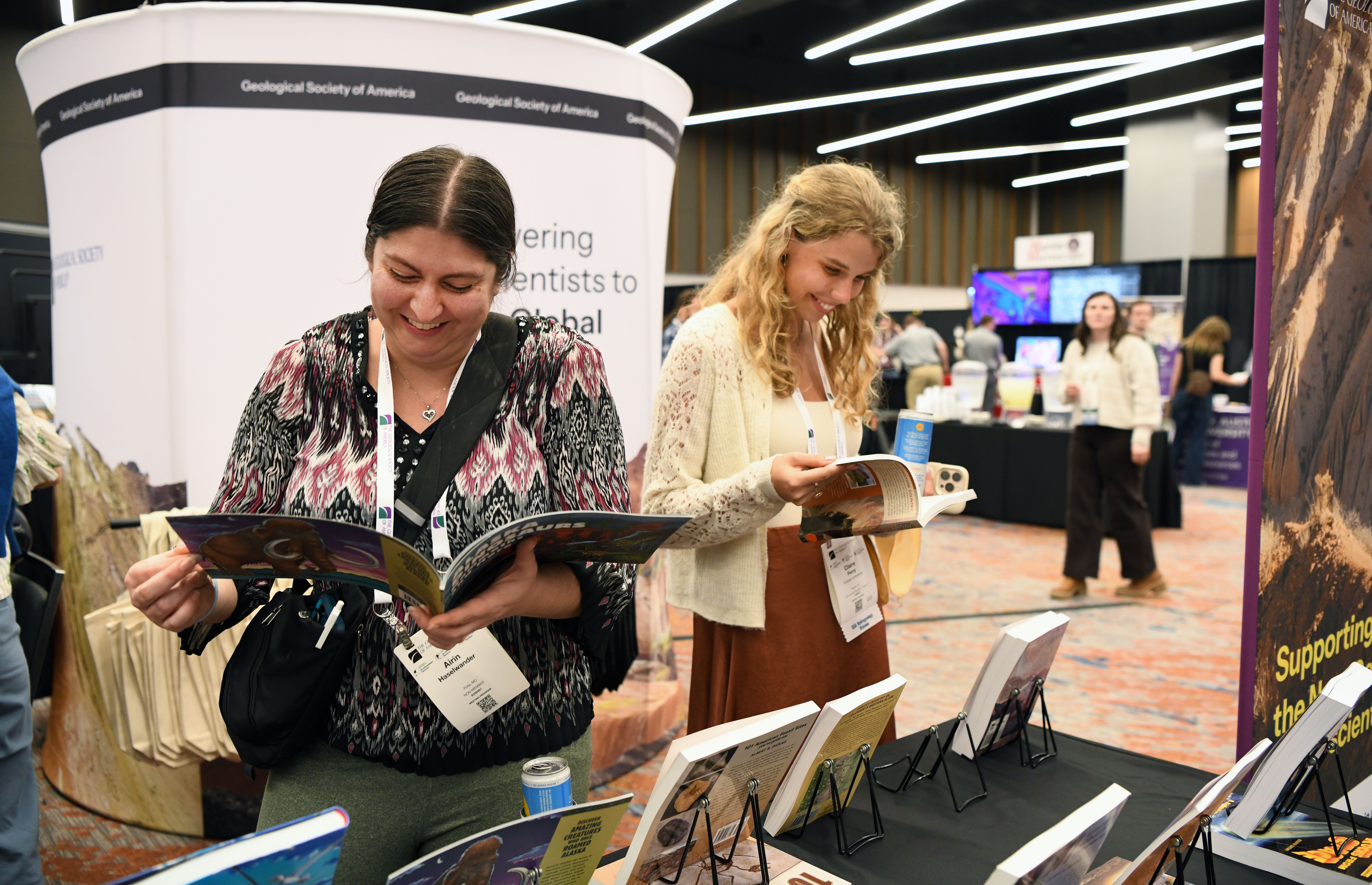 Two female GSA Triple Joint Section Meeting 2026 attendees wearing name badges smile while browsing publications at the GSA Store in the exhibit hall, with other exhibitors and attendees visible in the background.