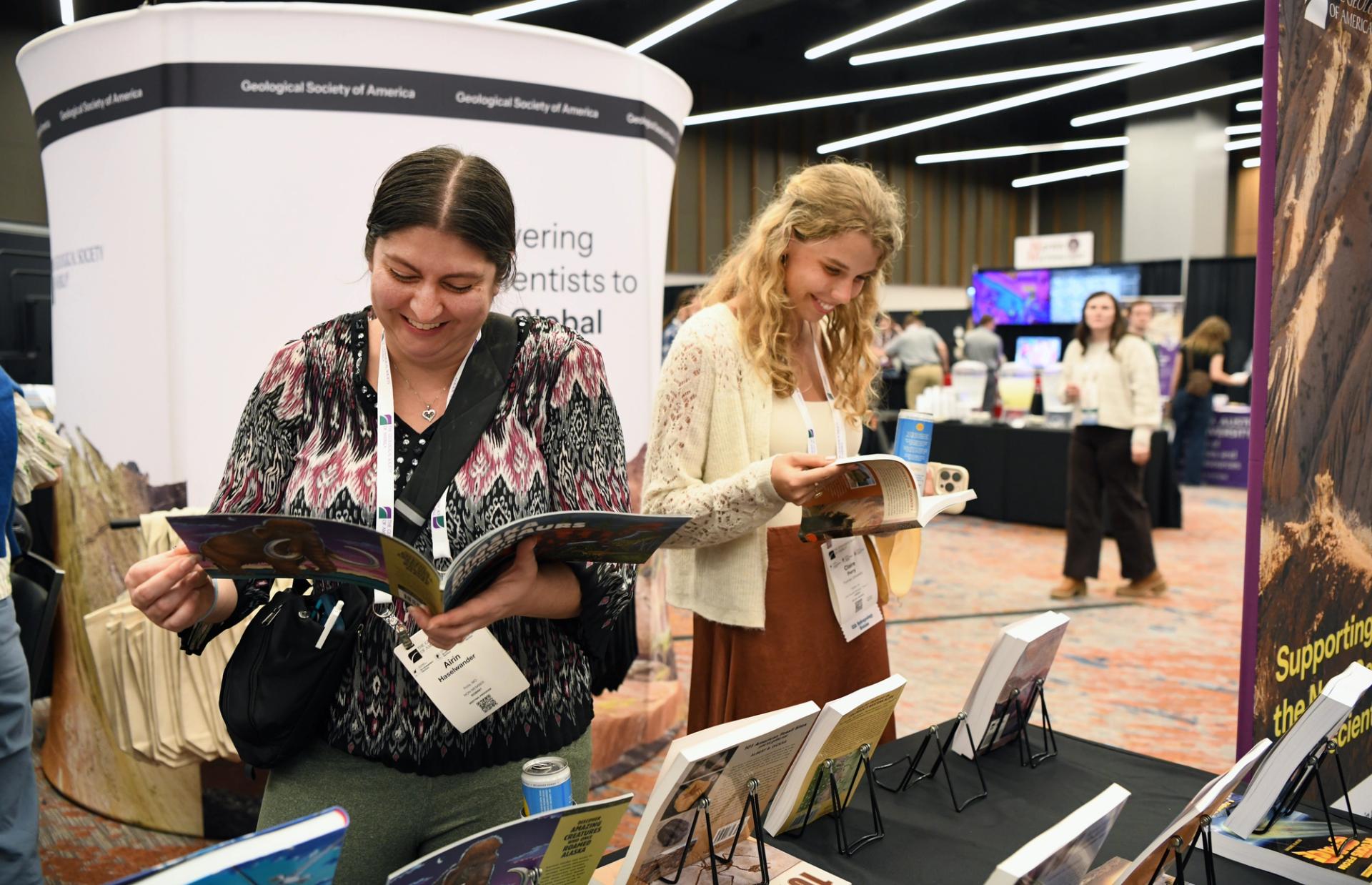 Two female GSA Triple Joint Section Meeting 2026 attendees wearing name badges smile while browsing publications at the GSA Store in the exhibit hall, with other exhibitors and attendees visible in the background.