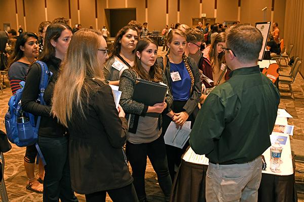 A group of people stands attentively around a booth at a career fair. They hold folders and papers, indicating interest and engagement. The setting is a large, well-lit room with a professional atmosphere.