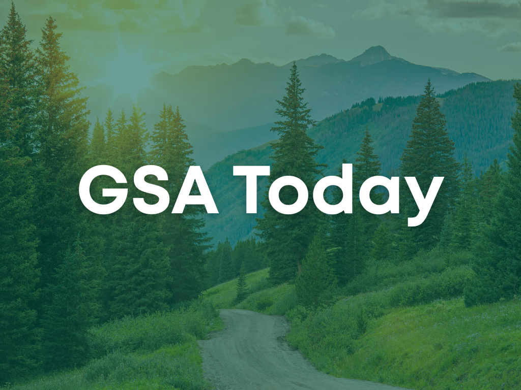 A sunlit, winding forest path surrounded by tall evergreen trees with distant mountains under blue skies. Text overlay reads "GSA Today."