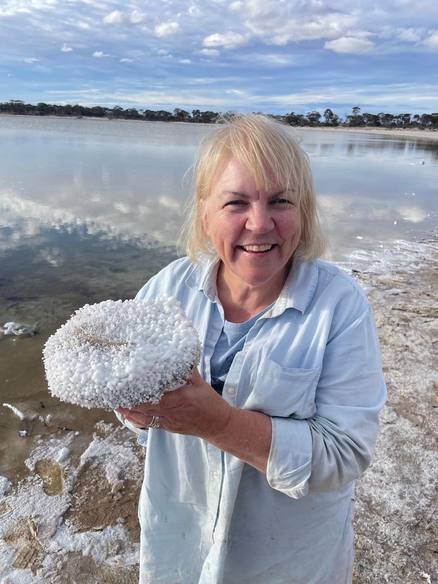 A blonde woman in a light blue shirt holds up a shell at the beach