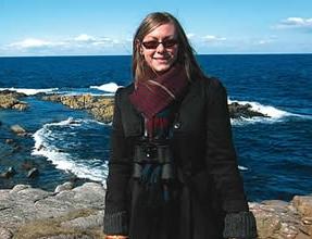 Tiffany Rivera, smiling, wearing a black coat, plaid scarf, and orange mittens, stands on a rocky coastal shoreline with binoculars around her neck, with deep blue ocean water and rocky outcrops visible behind her under a bright blue sky.