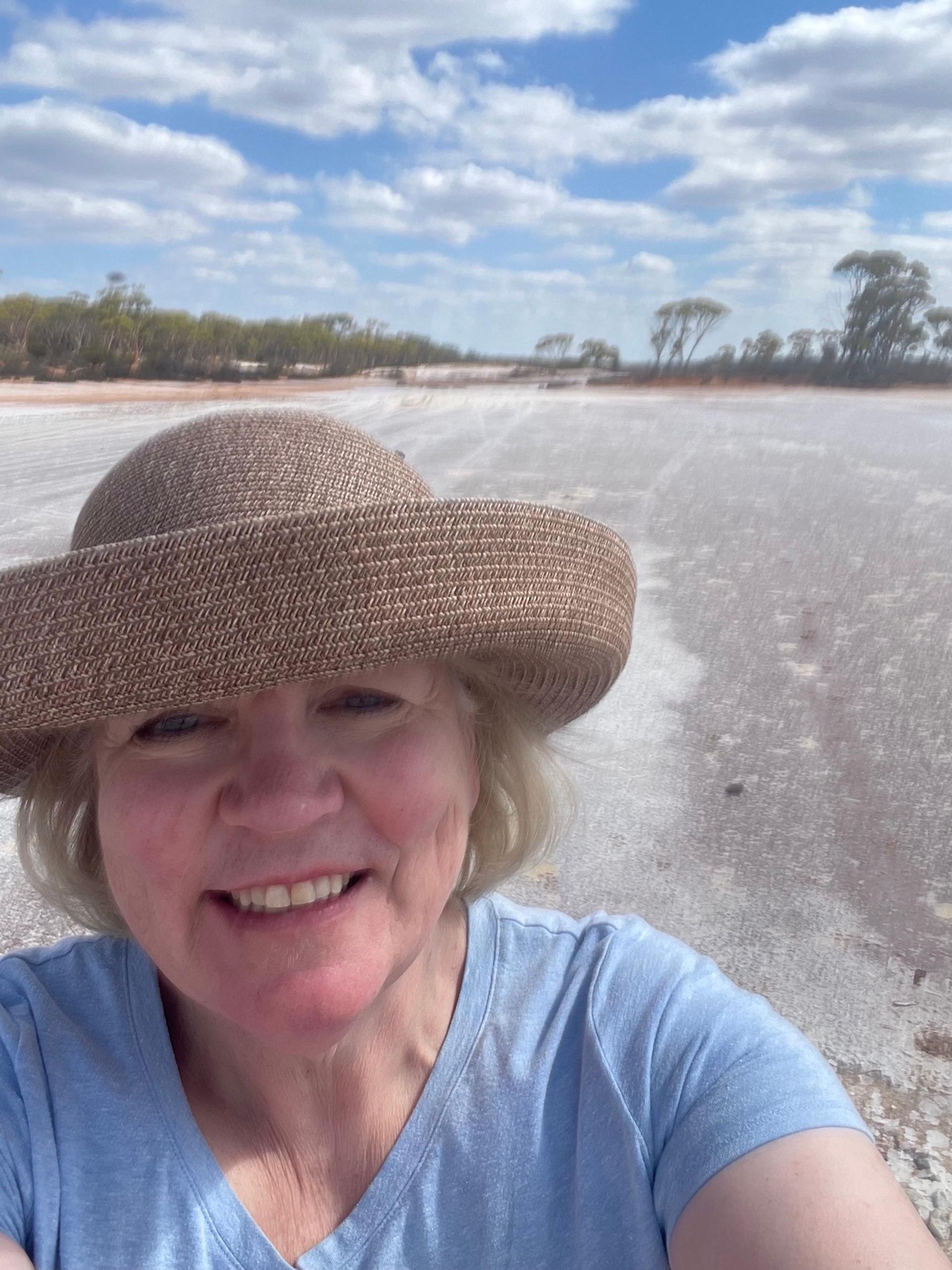 A woman in a light blue shirt and straw hat smiles while standing on the beach