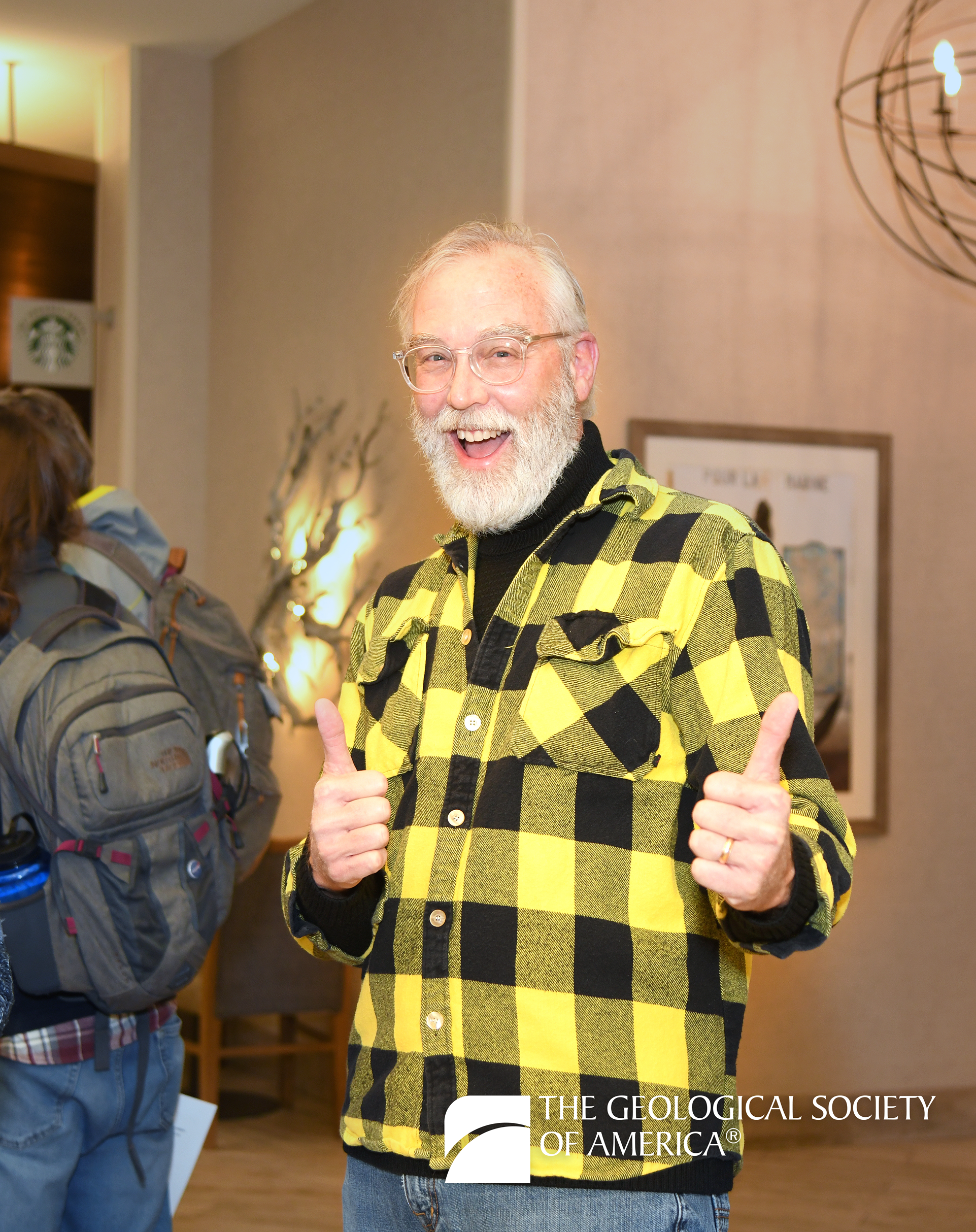 Elderly person in a yellow plaid shirt giving thumbs up, smiling energetically. Background shows a Starbucks and artwork. Geological Society logo visible.