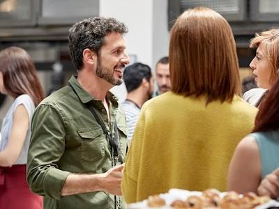 A group of people engaged in conversation at a social gathering. A man in a green shirt smiles while interacting with two women, creating a friendly atmosphere.