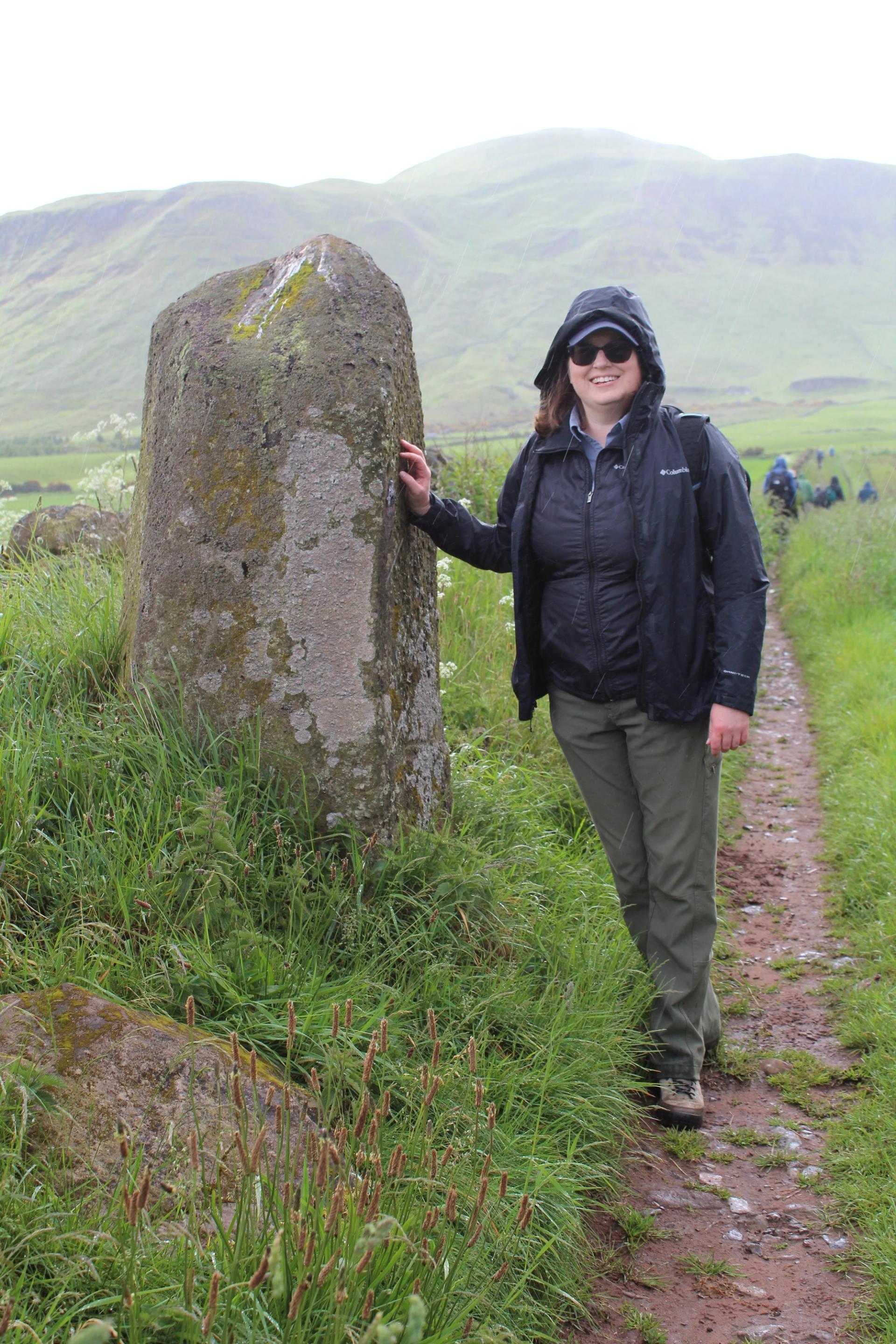 A woman in a black rain jacket and green pants stands on a misty trail and leans against a rock jutting out of the field.and points at a rock