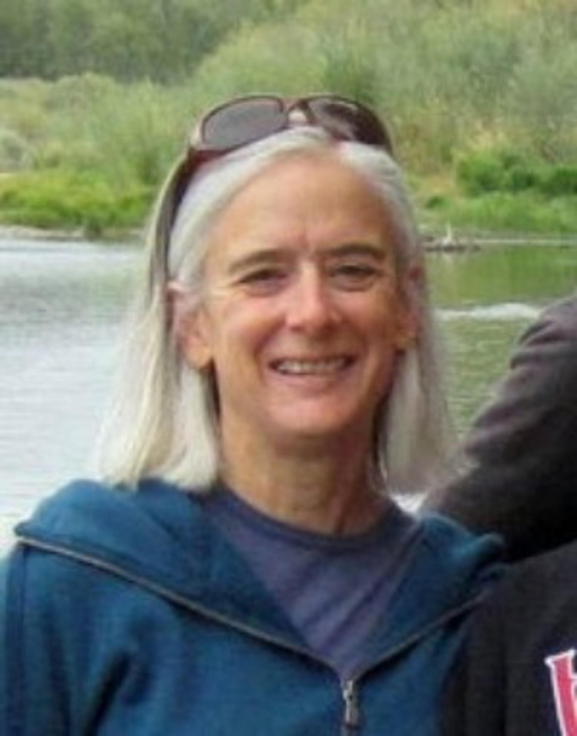 Nancy Riggs in a teal zippered shirt with water and green vegetation in the background.