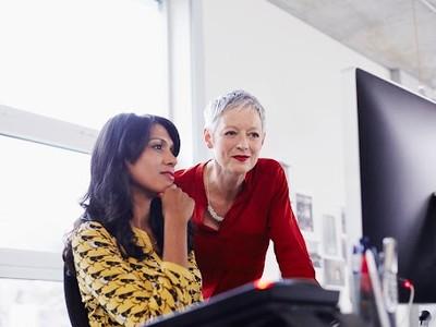 Two woman at a desk, one standing and one sitting looking at a computer.
