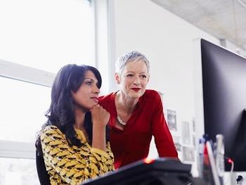 Two woman at a desk, one standing and one sitting looking at a computer.