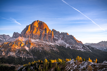 Warm sunlight bathes a rugged mountain peak, casting dramatic orange hues. Foreground features lush greenery under a clear blue sky with wispy clouds.