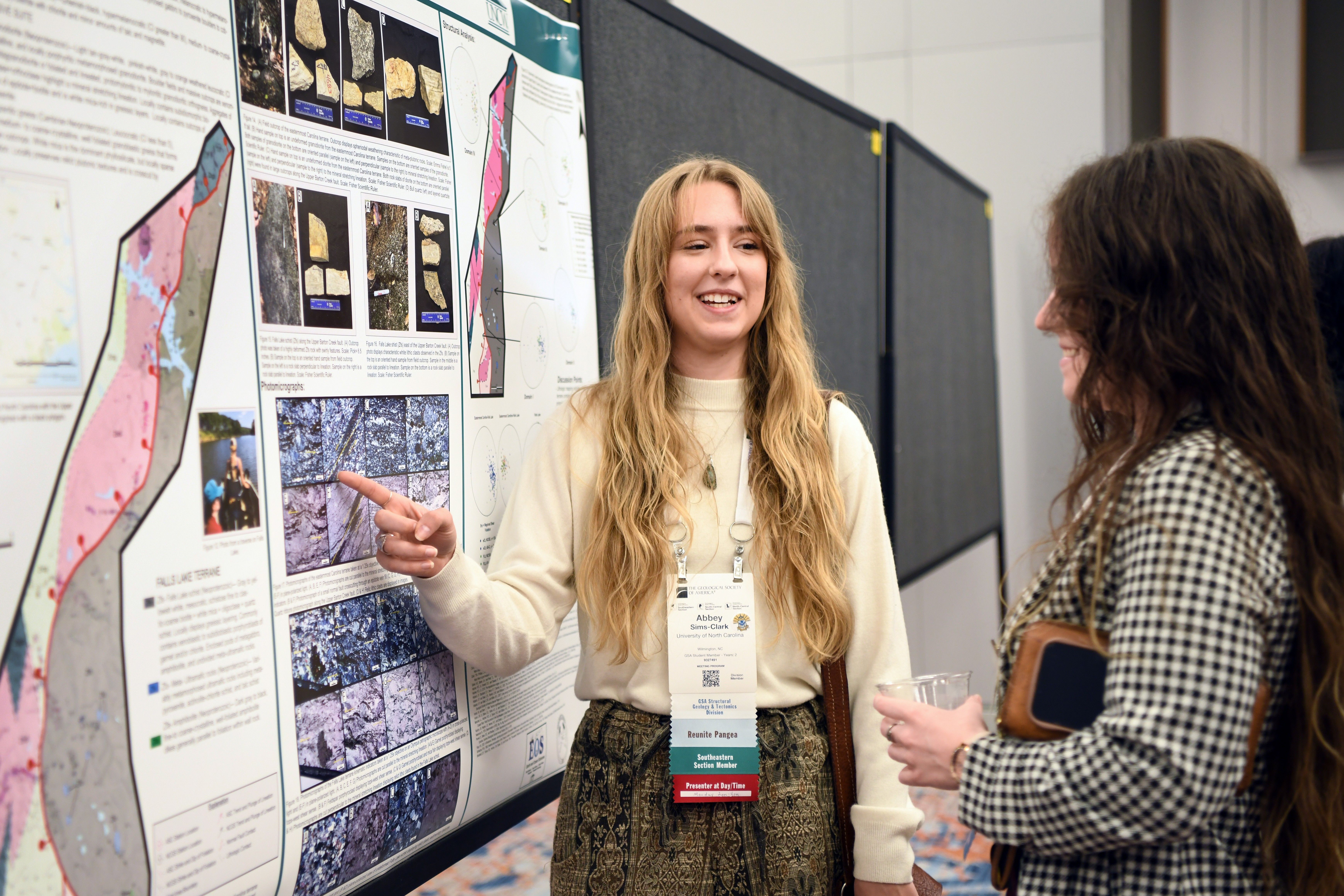 A young woman enthusiastically presents a scientific poster, gesturing at detailed maps and images, while another woman listens with interest.