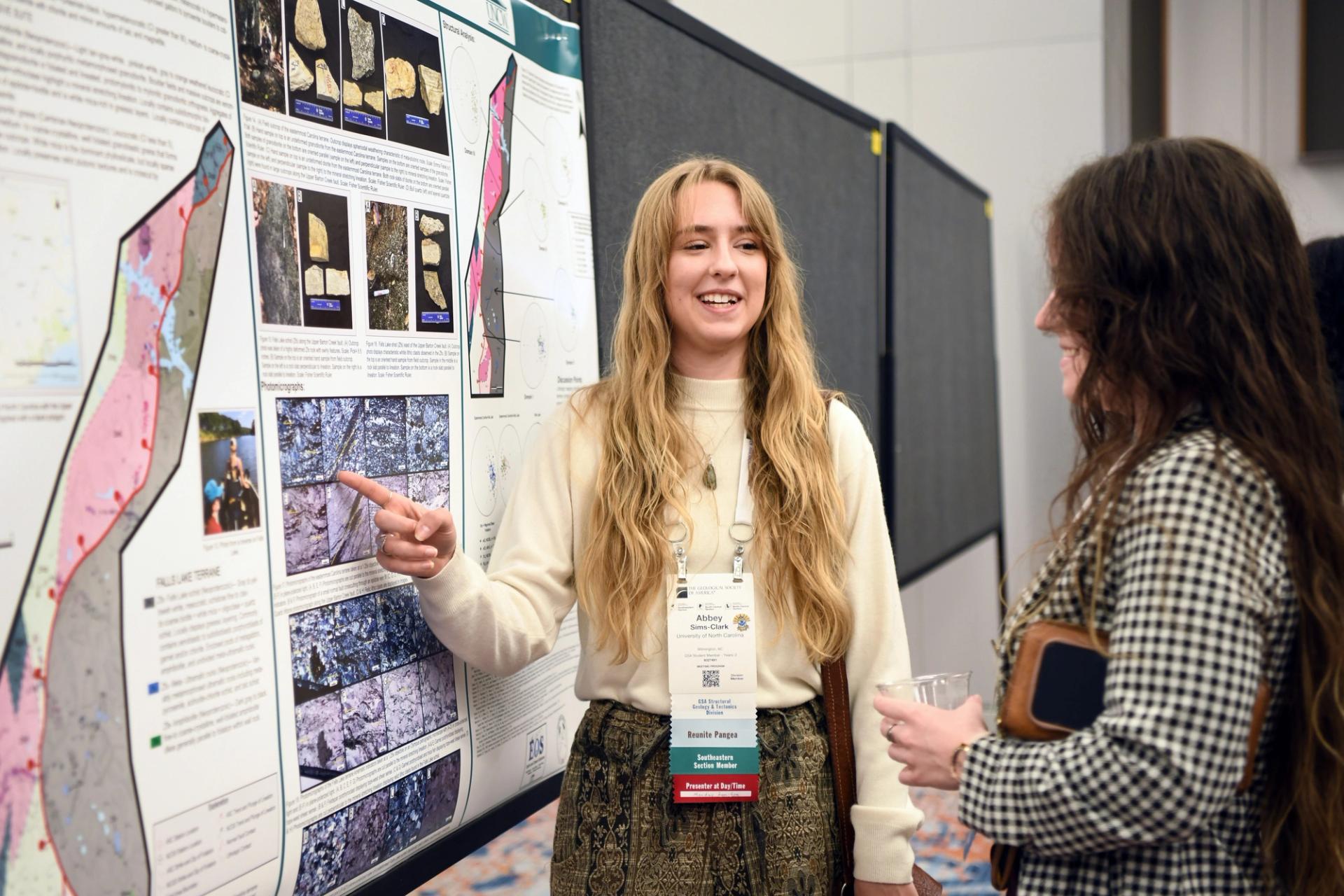 A young woman enthusiastically presents a scientific poster, gesturing at detailed maps and images, while another woman listens with interest.