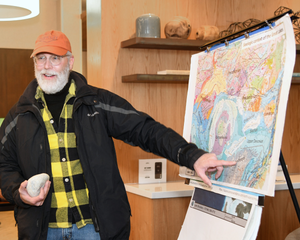 GSA instructor holding a rock, points at a colorful geological map on an easel. Background includes shelves with geological items.