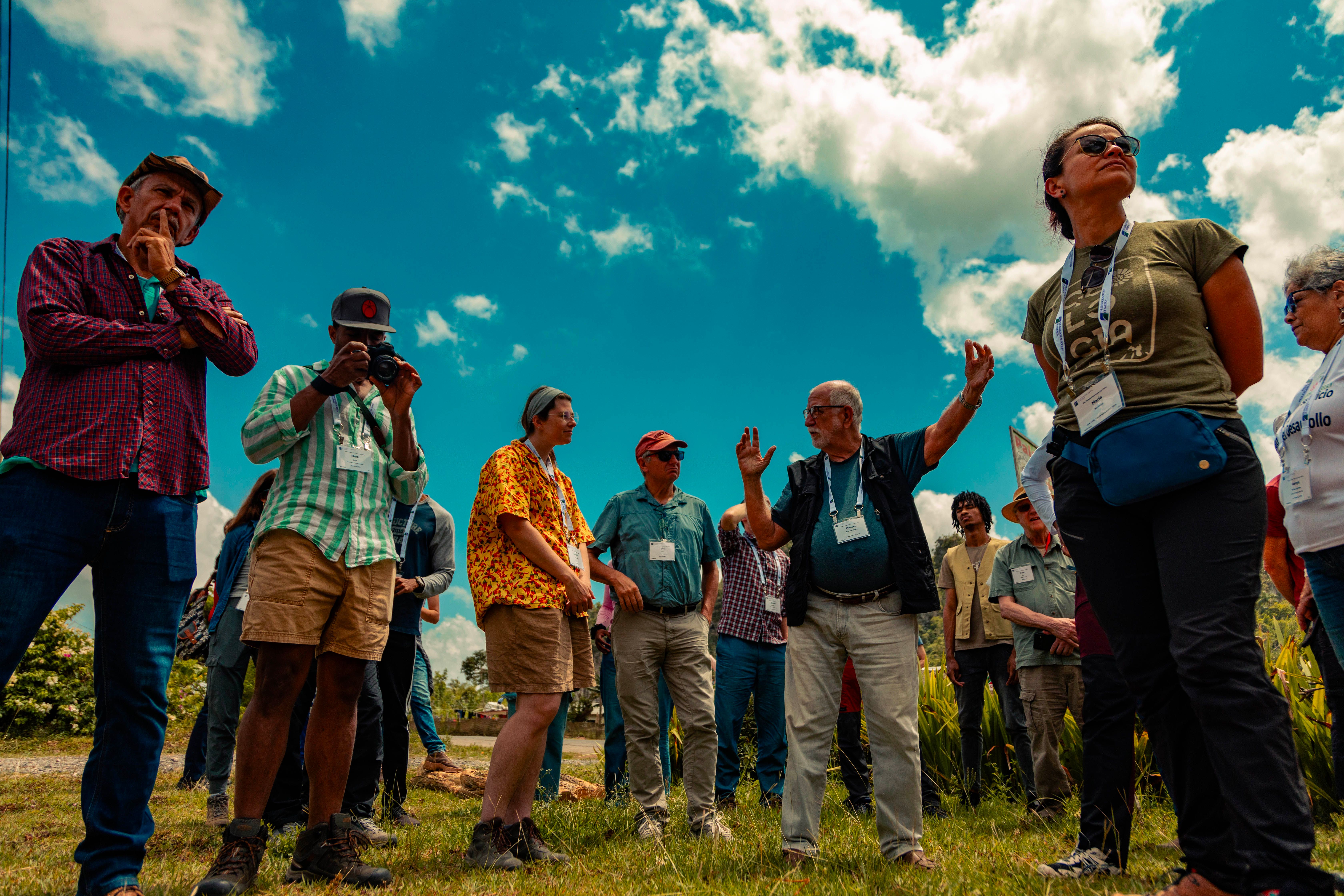 A group of GSA Thompson Field Forum 2025 participants wearing name badges listen attentively as a leader gestures and speaks during an outdoor field session under a bright blue sky in Viñales, Cuba.