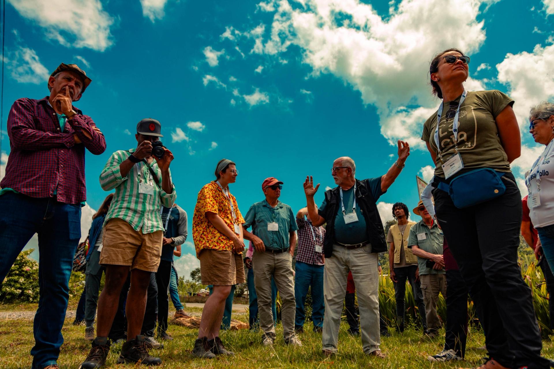A group of GSA Thompson Field Forum 2025 participants wearing name badges listen attentively as a leader gestures and speaks during an outdoor field session under a bright blue sky in Viñales, Cuba.