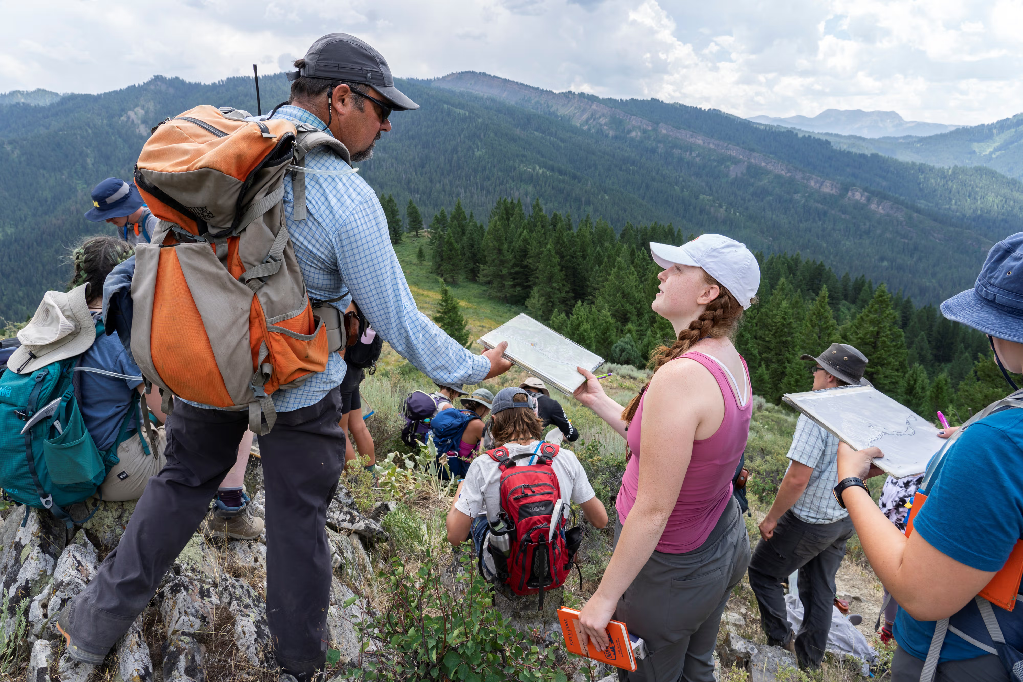 A group of students and an instructor in hiking gear stand on a rocky mountain ridge, examining maps and notebooks against a backdrop of forested hills.