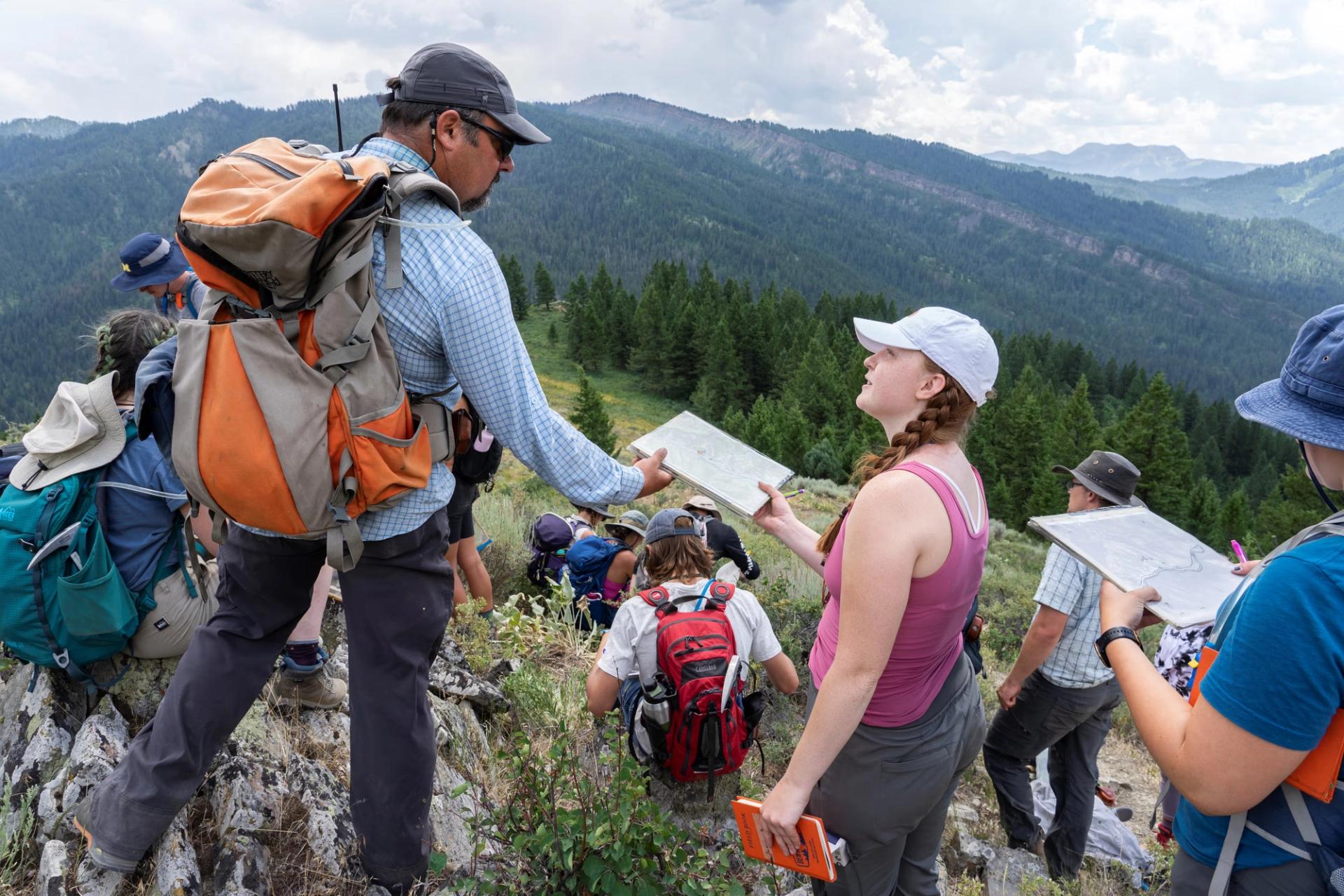 A group of students and an instructor in hiking gear stand on a rocky mountain ridge, examining maps and notebooks against a backdrop of forested hills.