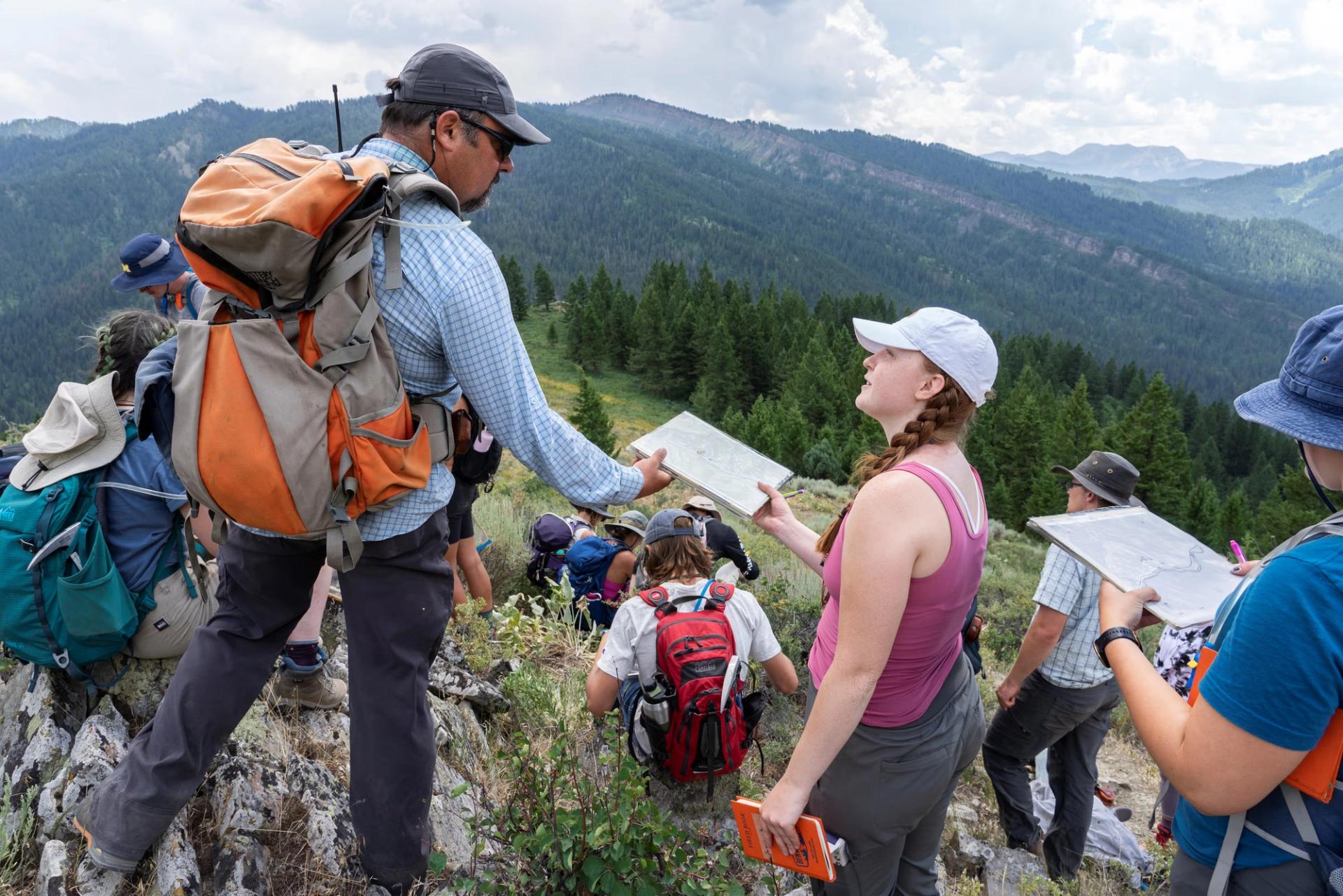 A group of students and an instructor in hiking gear stand on a rocky mountain ridge, examining maps and notebooks against a backdrop of forested hills.