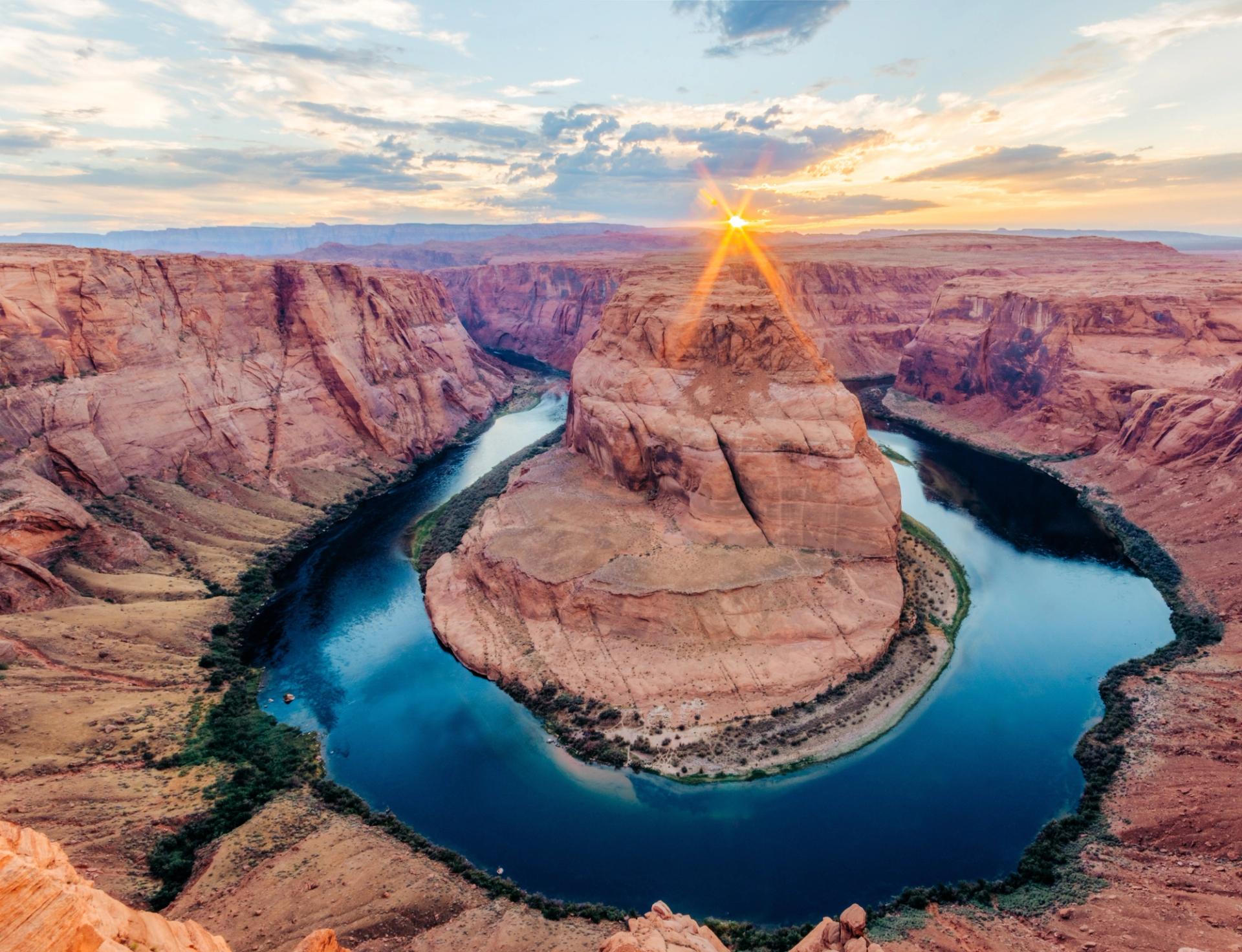 An aerial view of Horseshoe Bend near Page, Arizona, showing the Colorado River wrapping in a dramatic horseshoe curve around a towering sandstone butte, with vibrant red canyon walls and a stunning sunburst sunset illuminating the sky above.