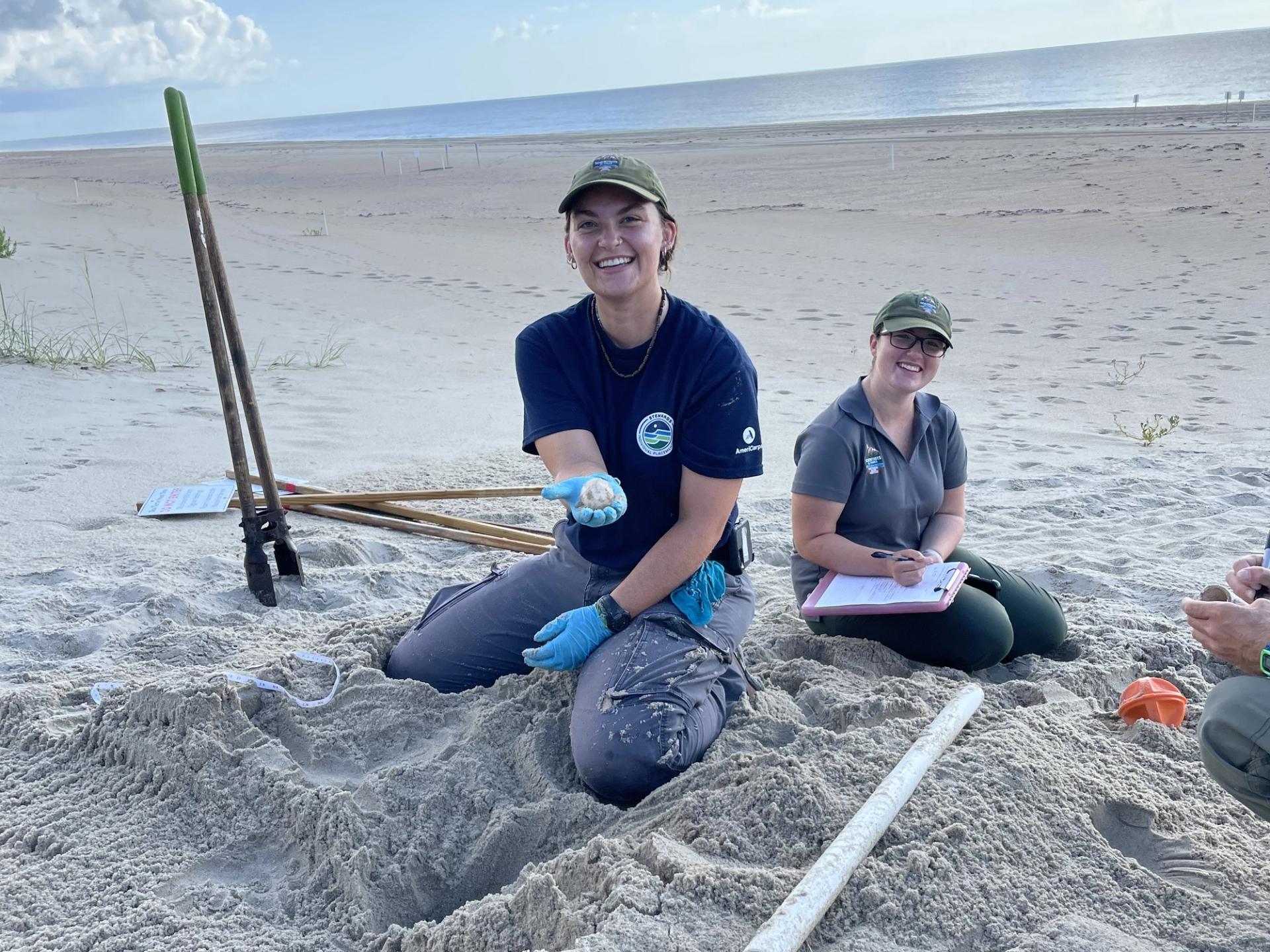 Photo of Scientists in the Parks Participants in the field on a beach sitting in sand holding a sample up to the camera