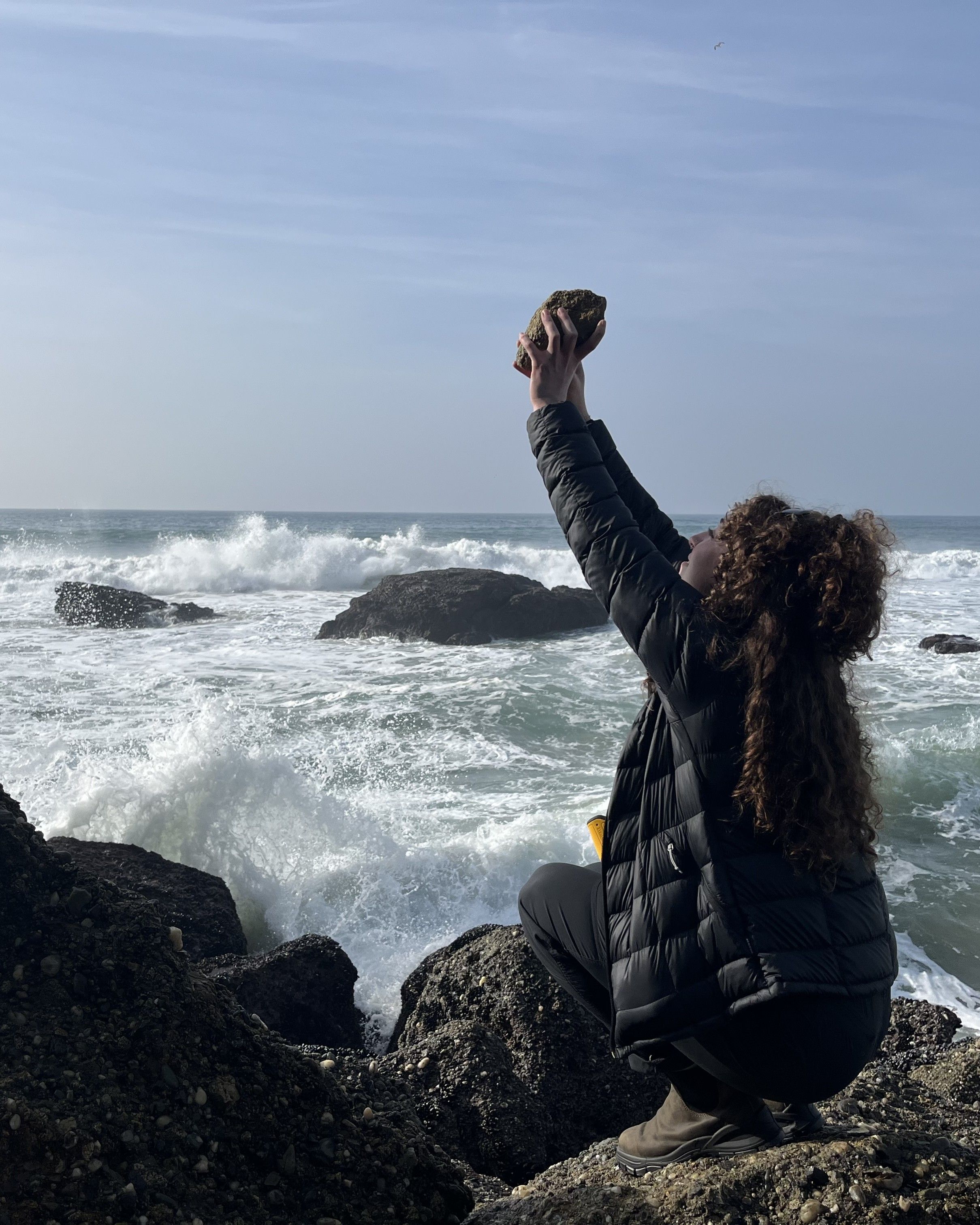 Photo of GSA Graduate Student Research Grant Recipient conducting fieldwork holding rock up to the sky on a rocky shoreline
