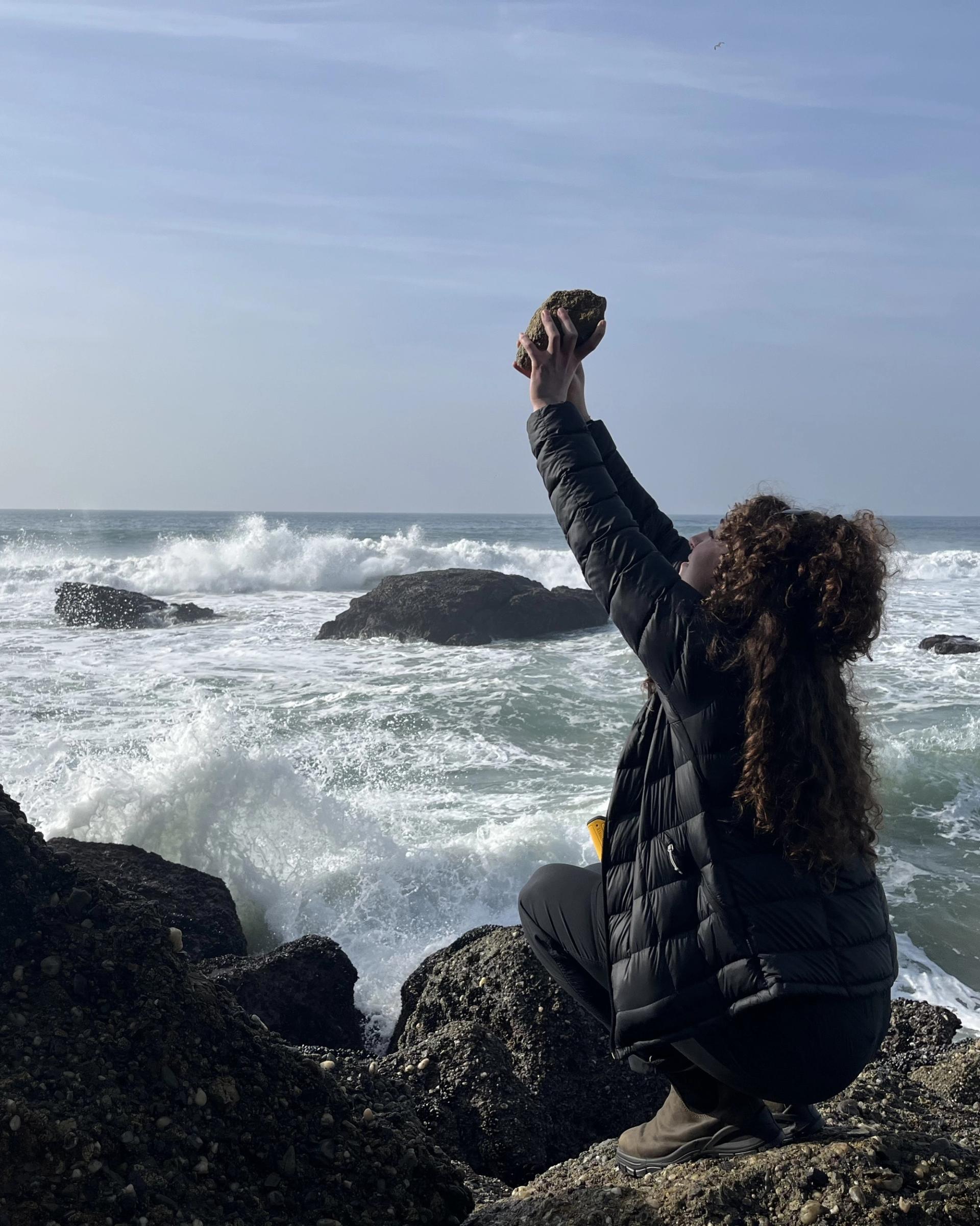 Photo of GSA Graduate Student Research Grant Recipient conducting fieldwork holding rock up to the sky on a rocky shoreline