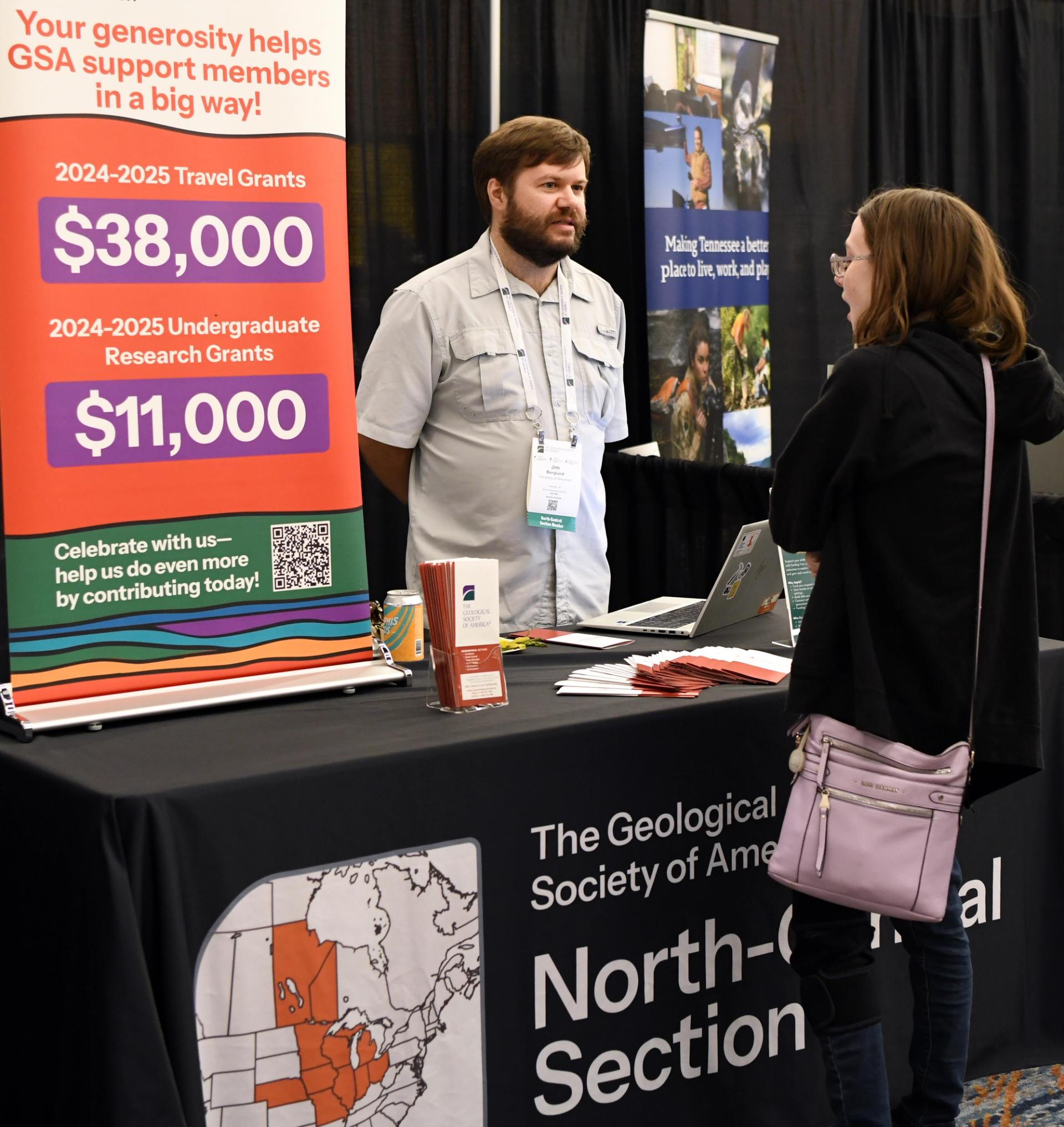 A man and woman converse at a Geological Society of America booth. A sign displays grant amounts. The booth is informative and professional.