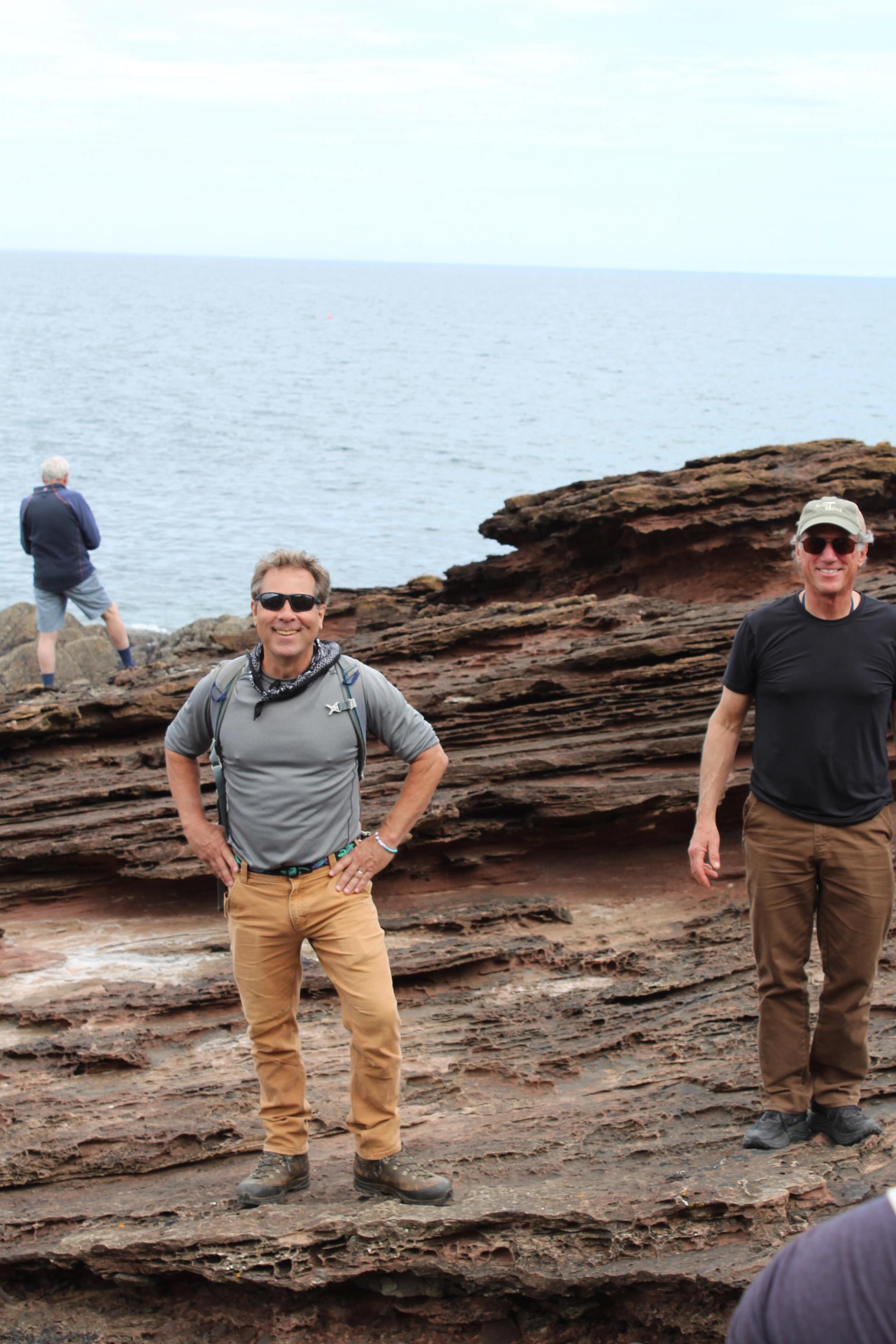 Two men in outdoor gear and sunglasses stand atop a striated rock formation with a body of water in the background.
