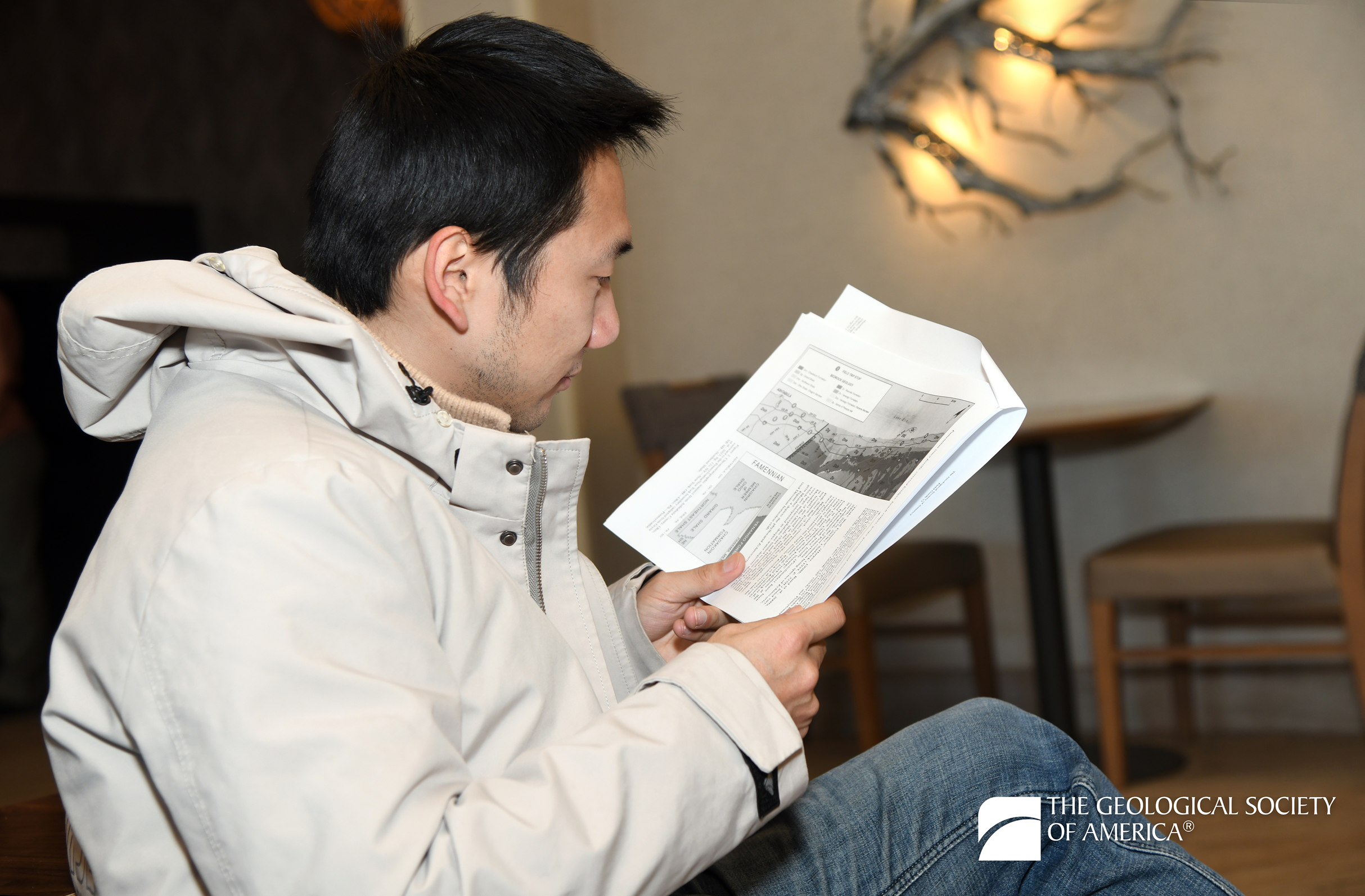 A person in a beige coat sits reading a scientific paper. The background features a modern art piece and soft lighting. The atmosphere is calm and studious.