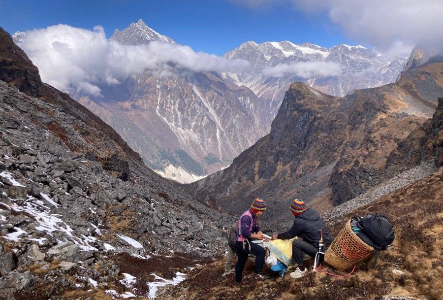 Two people in colorful hats sit on a rocky mountain pass, surrounded by snow-dusted peaks and a clear blue sky, conveying adventure and tranquility.