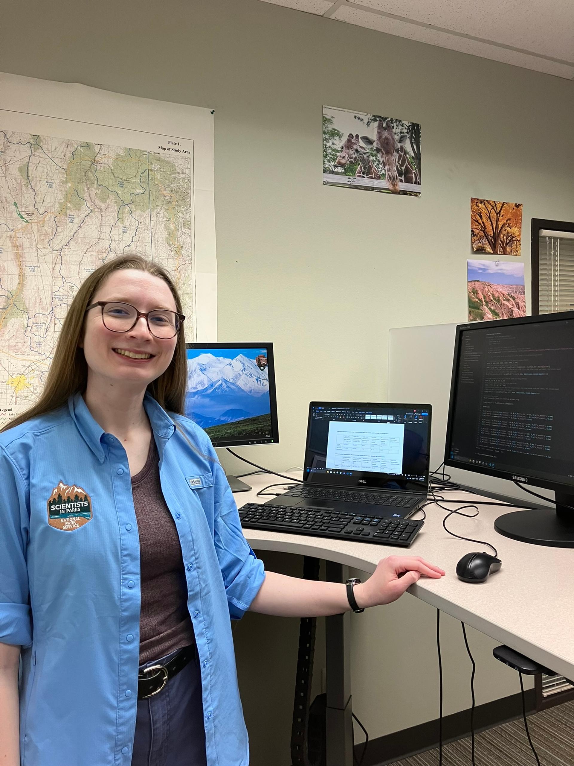 A smiling woman wearing a blue "Scientists in Parks" shirt stands at a desk with multiple monitors displaying data and code. A large topographic map and nature photos are pinned to the wall behind her.