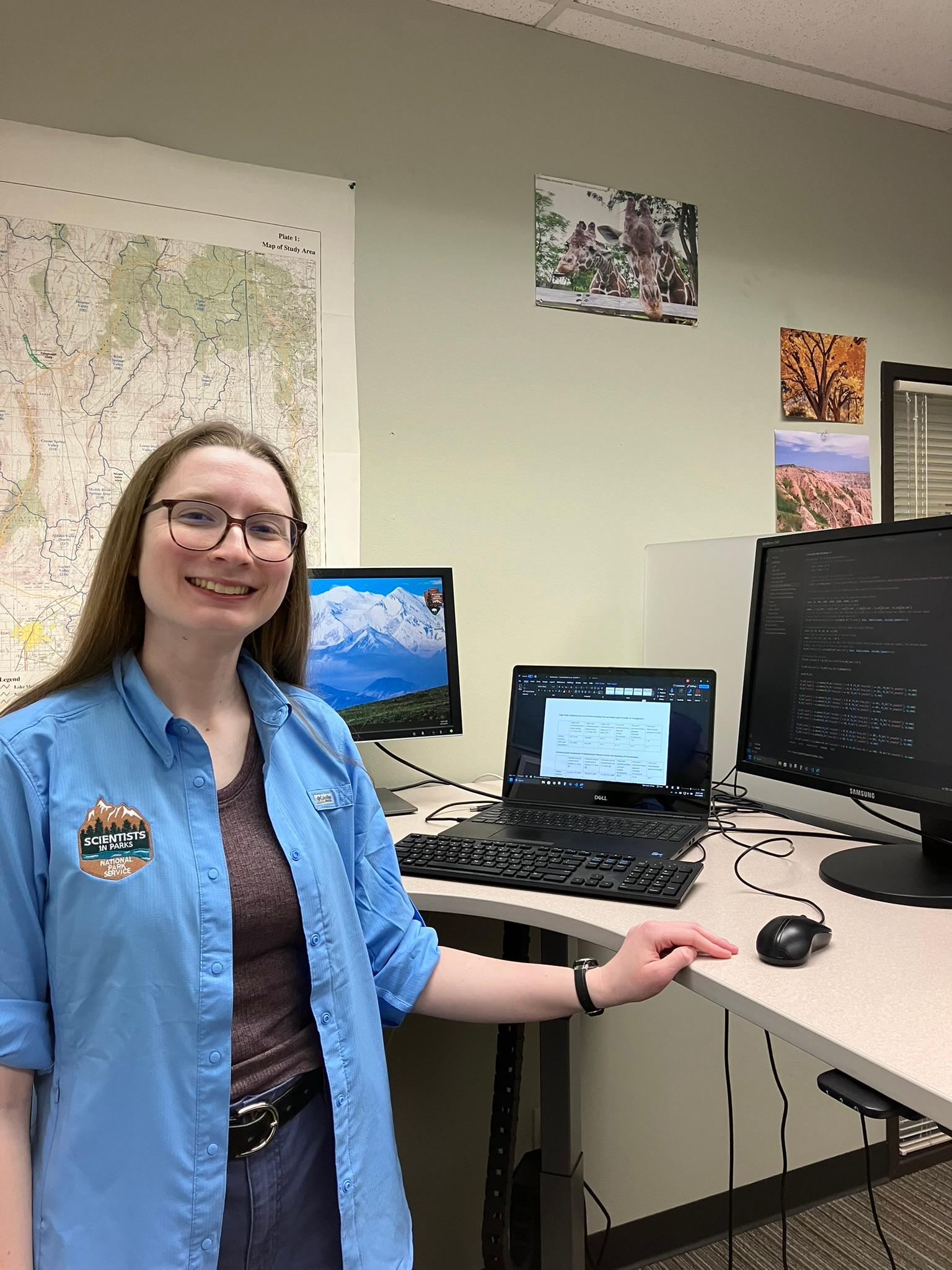 A smiling woman wearing a blue "Scientists in Parks" shirt stands at a desk with multiple monitors displaying data and code. A large topographic map and nature photos are pinned to the wall behind her.