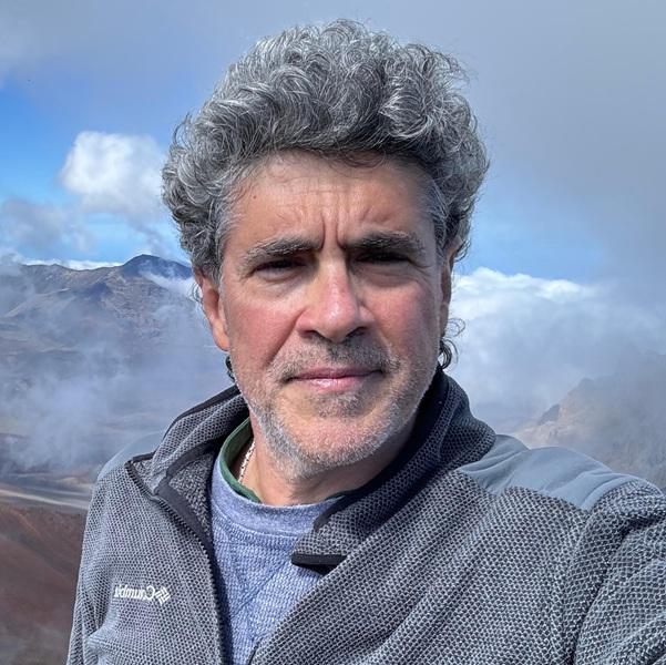 Valerio Acocella in a gray jacket and blue shirt with mountains, clouds, and blue sky in the background.