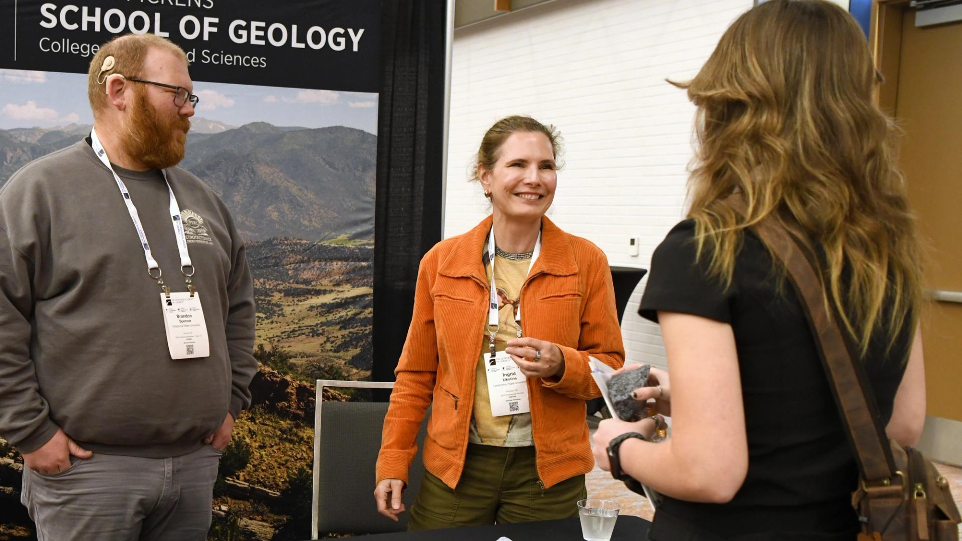 Three people engage in conversation at a geology school exhibit. A woman in an orange jacket holds a rock sample, smiling warmly. Keywords include geology, education, and interaction.