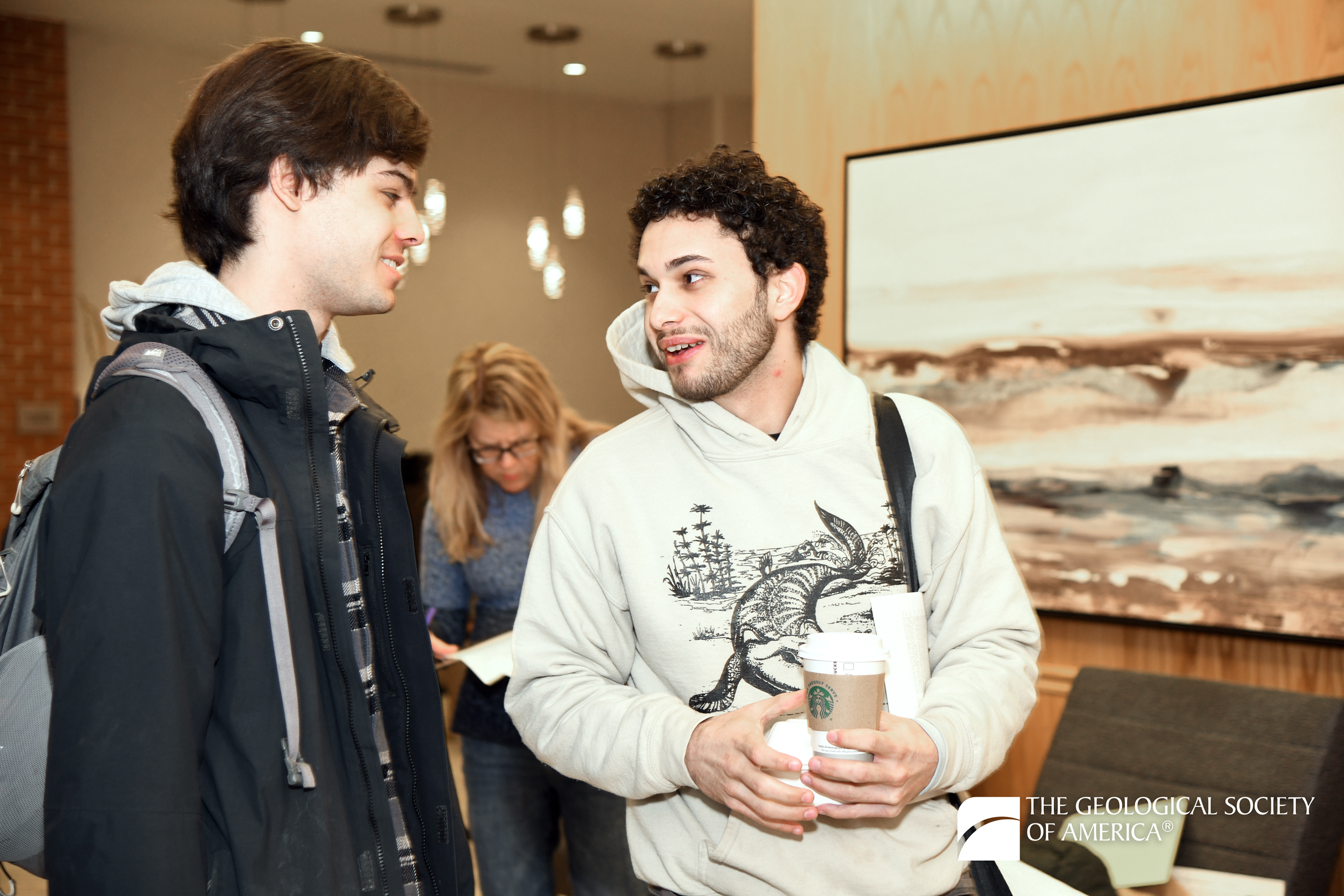 Two people in casual attire, holding coffee cups, are engaged in a friendly conversation indoors. A third person is in the background, focused on reading. The mood is relaxed, with a painting and the Geological Society of America logo visible.