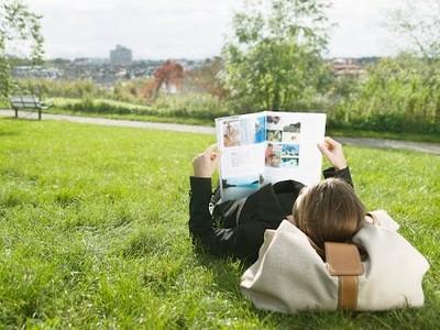 A person lies on grass in a park, reading a magazine with colorful pages. A bag serves as a pillow. The scene is sunny and serene, with a blurred city skyline.