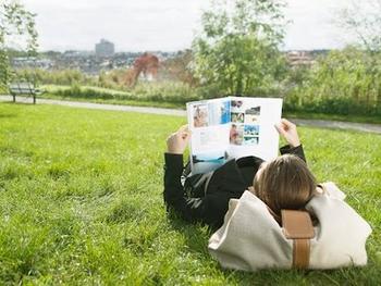 A person lies on grass in a park, reading a magazine with colorful pages. A bag serves as a pillow. The scene is sunny and serene, with a blurred city skyline.