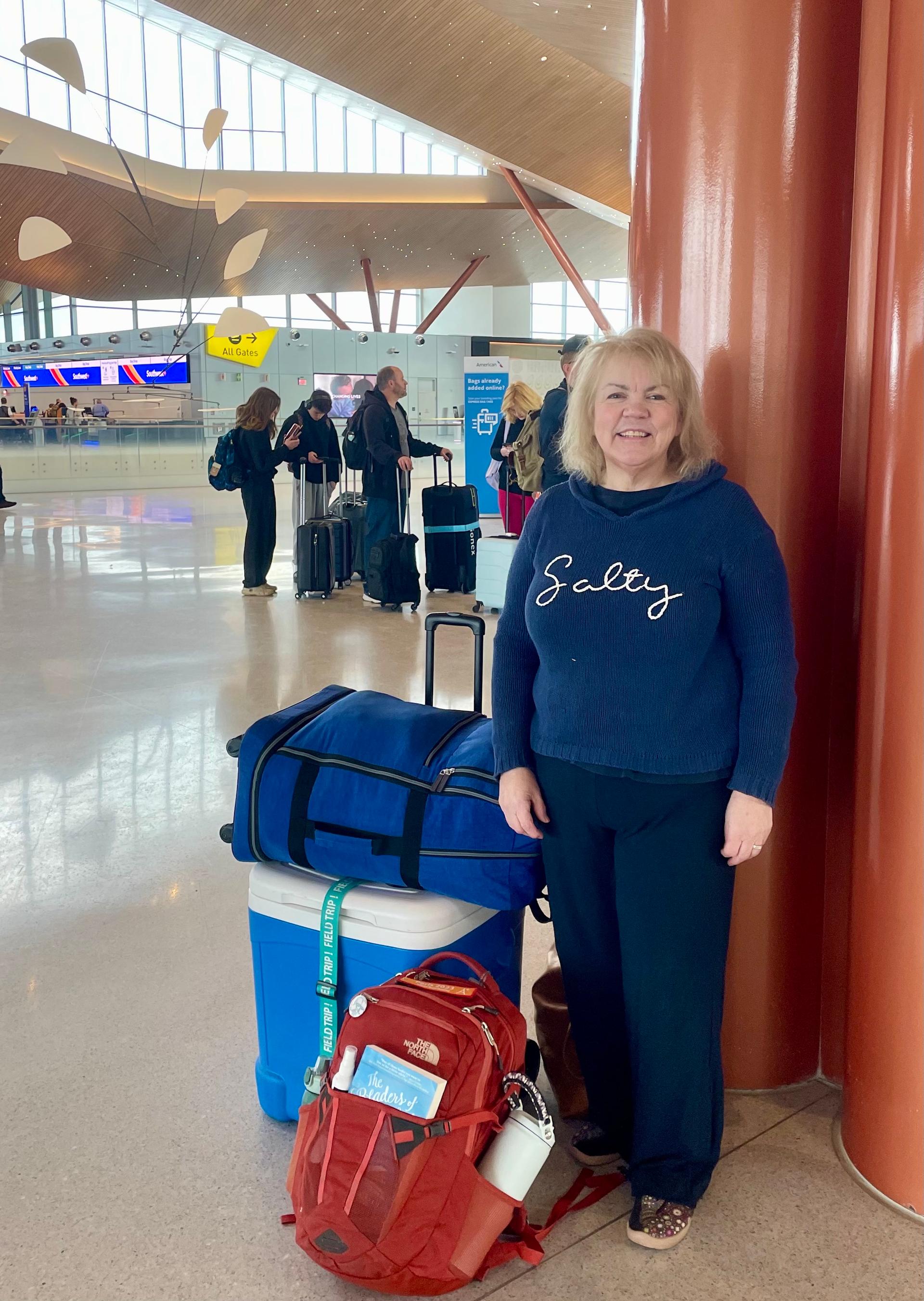 A woman in a navy shirt and black pants stands next to luggage at the airport.