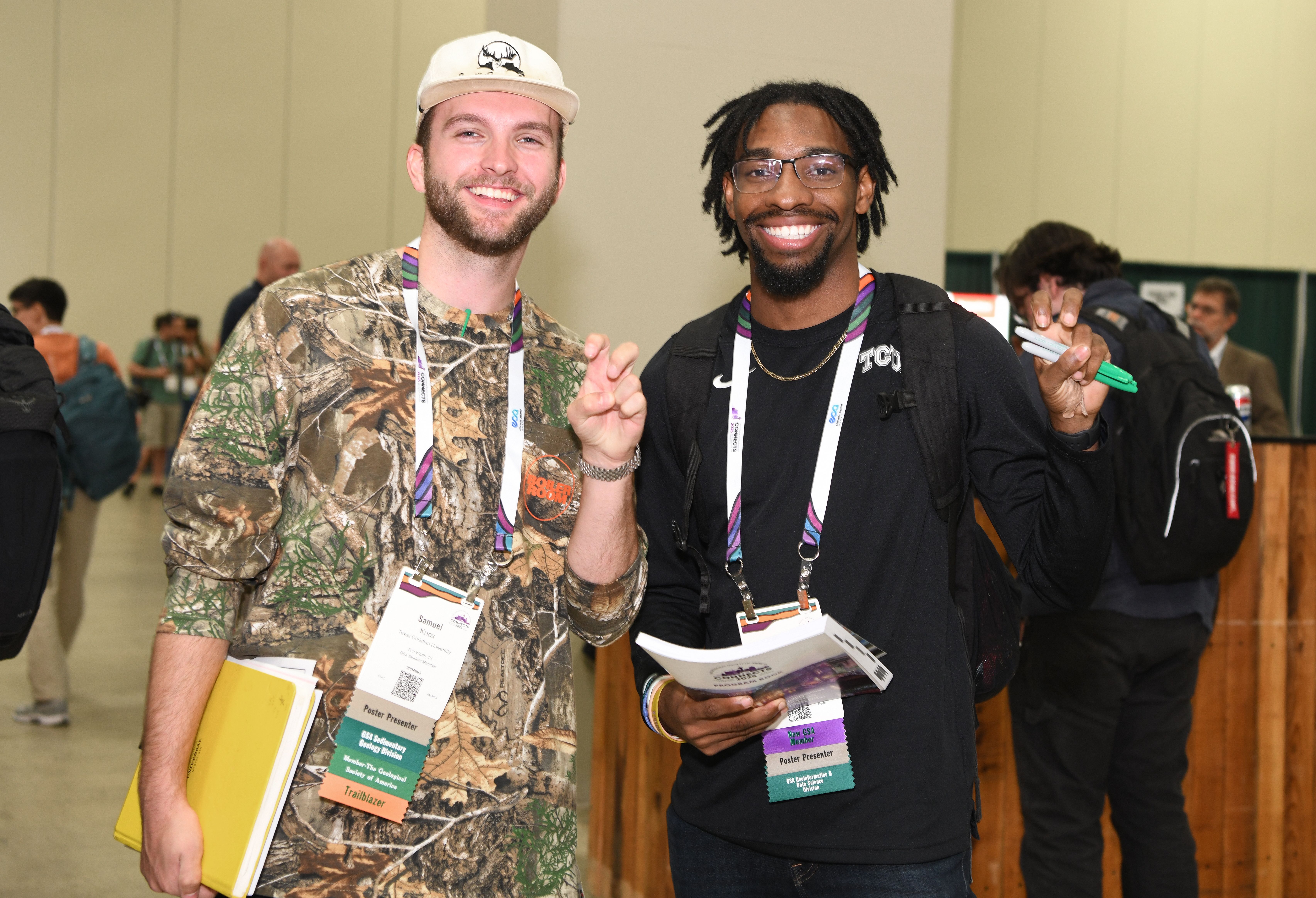 Two smiling young male GSA conference attendees wearing poster presenter badges and colorful lanyards pose together in a busy conference hall, one holding a yellow folder and the other holding conference materials.