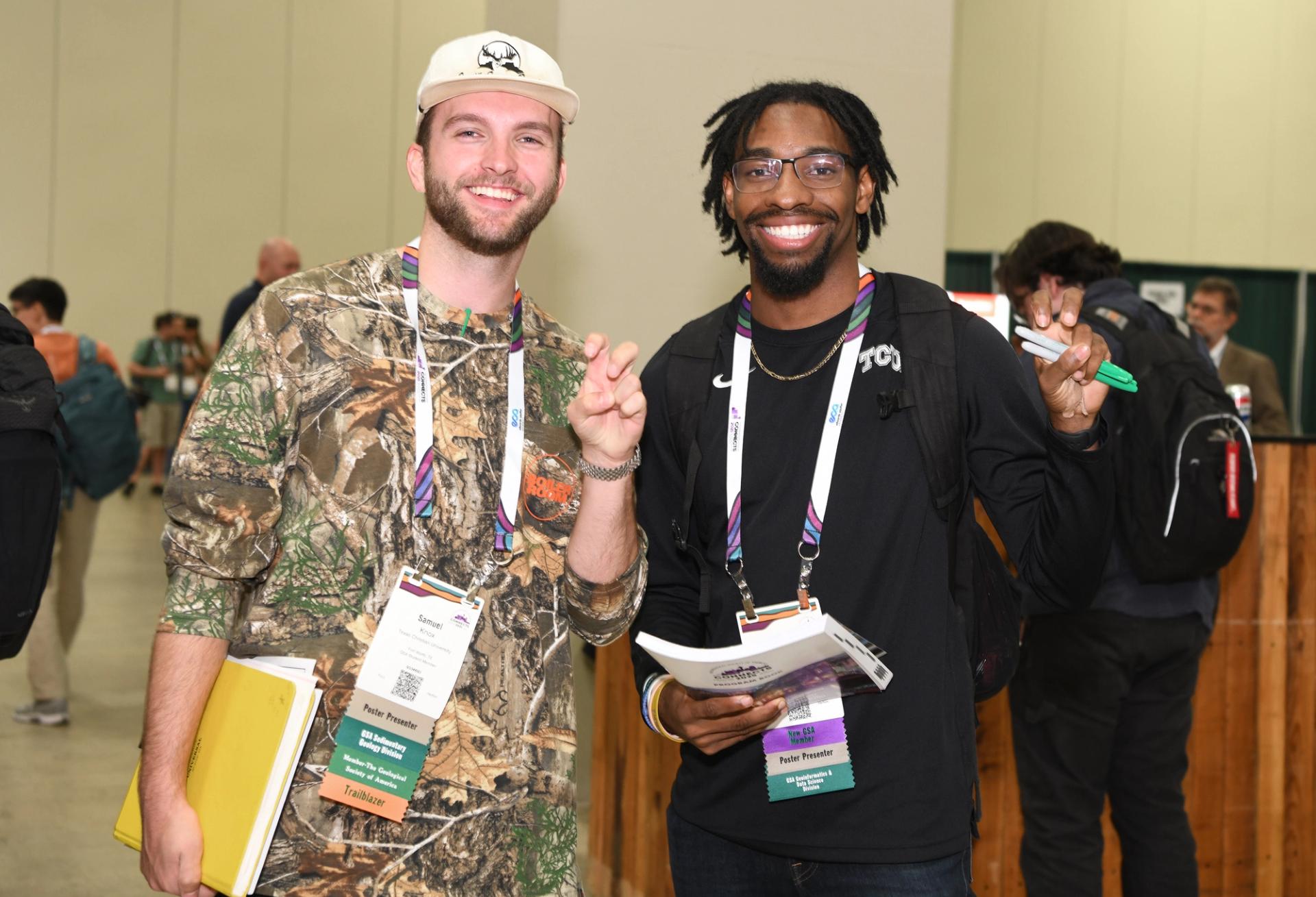 Two smiling young male GSA conference attendees wearing poster presenter badges and colorful lanyards pose together in a busy conference hall, one holding a yellow folder and the other holding conference materials.