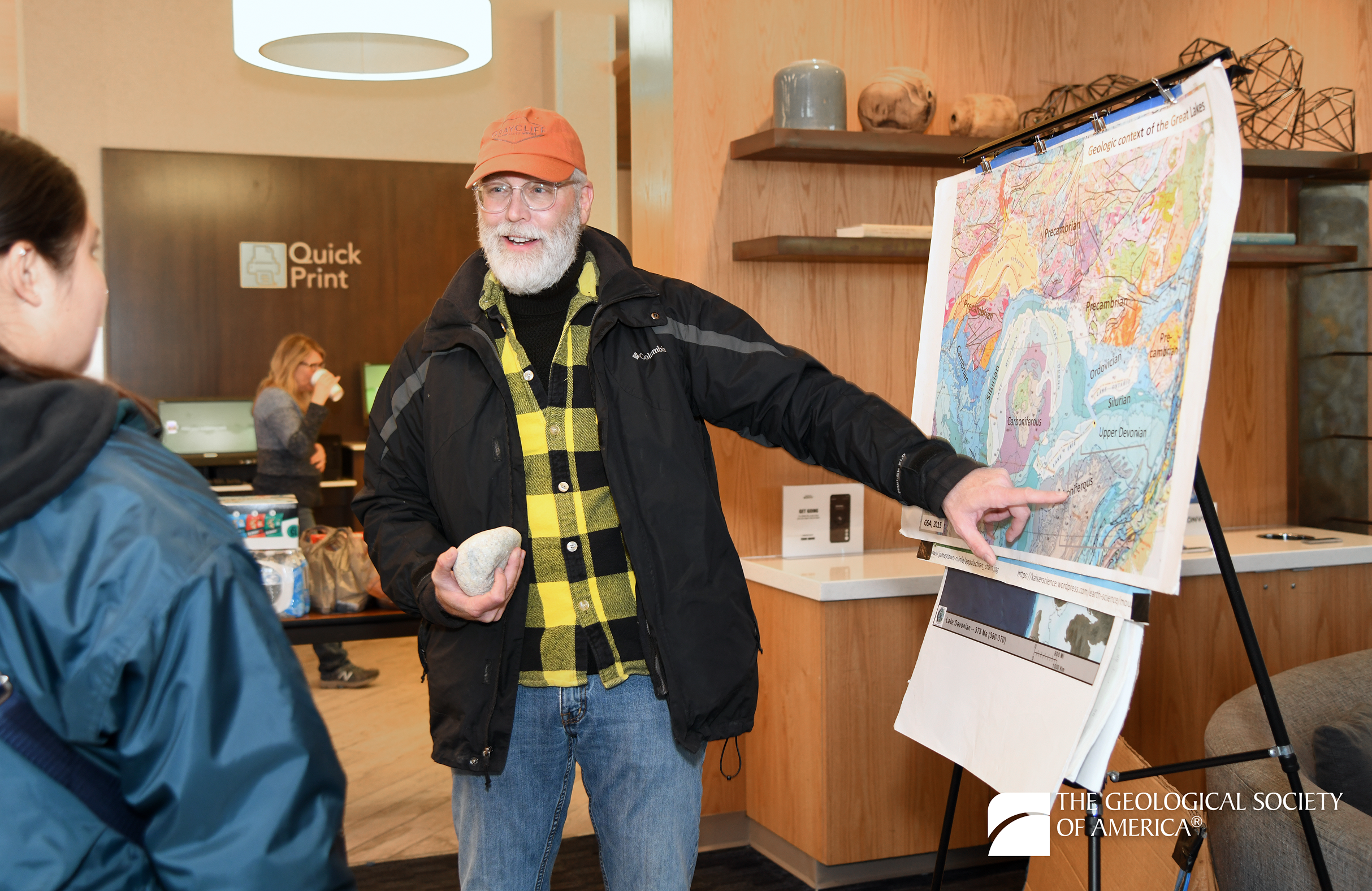 A man in a plaid shirt and cap enthusiastically gestures to a colorful geological map on an easel, holding a rock, engaging with a woman in the foreground.