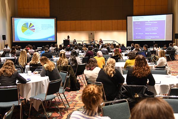 Audience sits at round tables in a conference room, listening to a speaker with a presentation projected on screens. The atmosphere is focused and attentive.