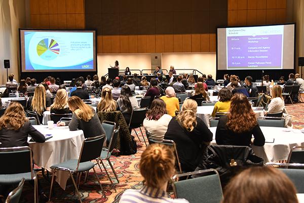 Audience sits at round tables in a conference room, listening to a speaker with a presentation projected on screens. The atmosphere is focused and attentive.