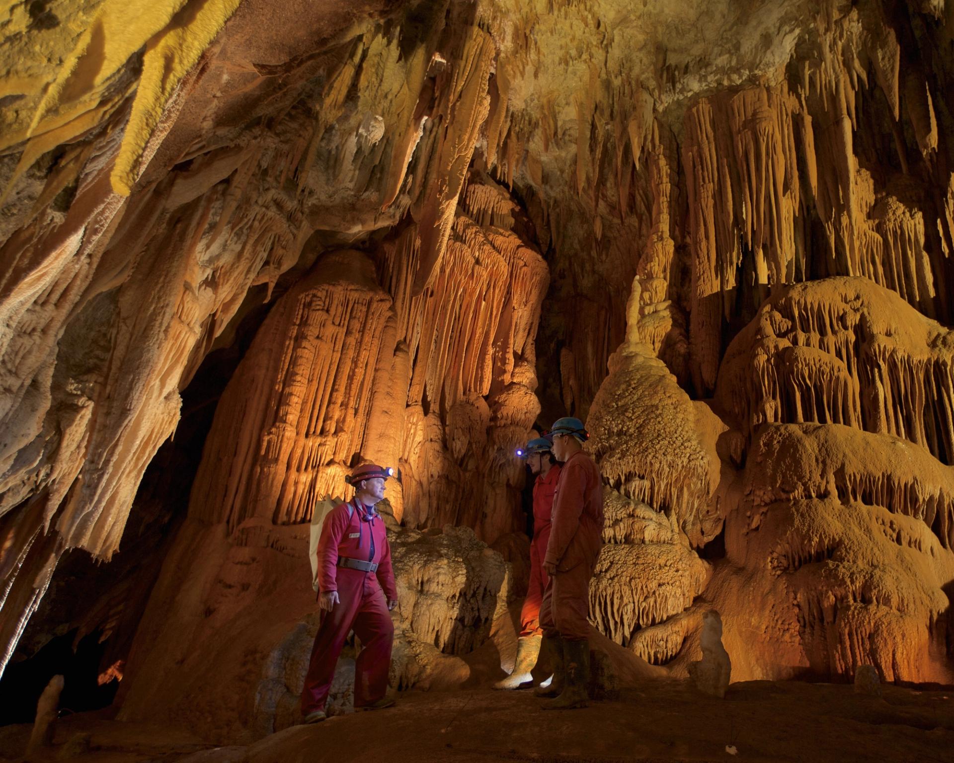 Three GSA mambers in red overalls and helmets with headlamps stand in a large cave, surrounded by stunning stalactites and stalagmites, exuding awe.