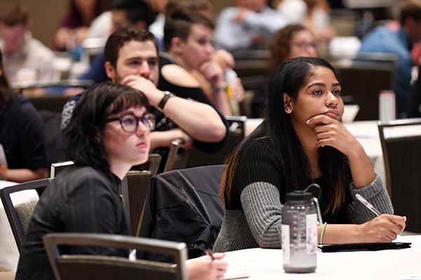 A diverse group of people sit at tables in a conference room, attentively listening and taking notes. The atmosphere is focused and engaged.